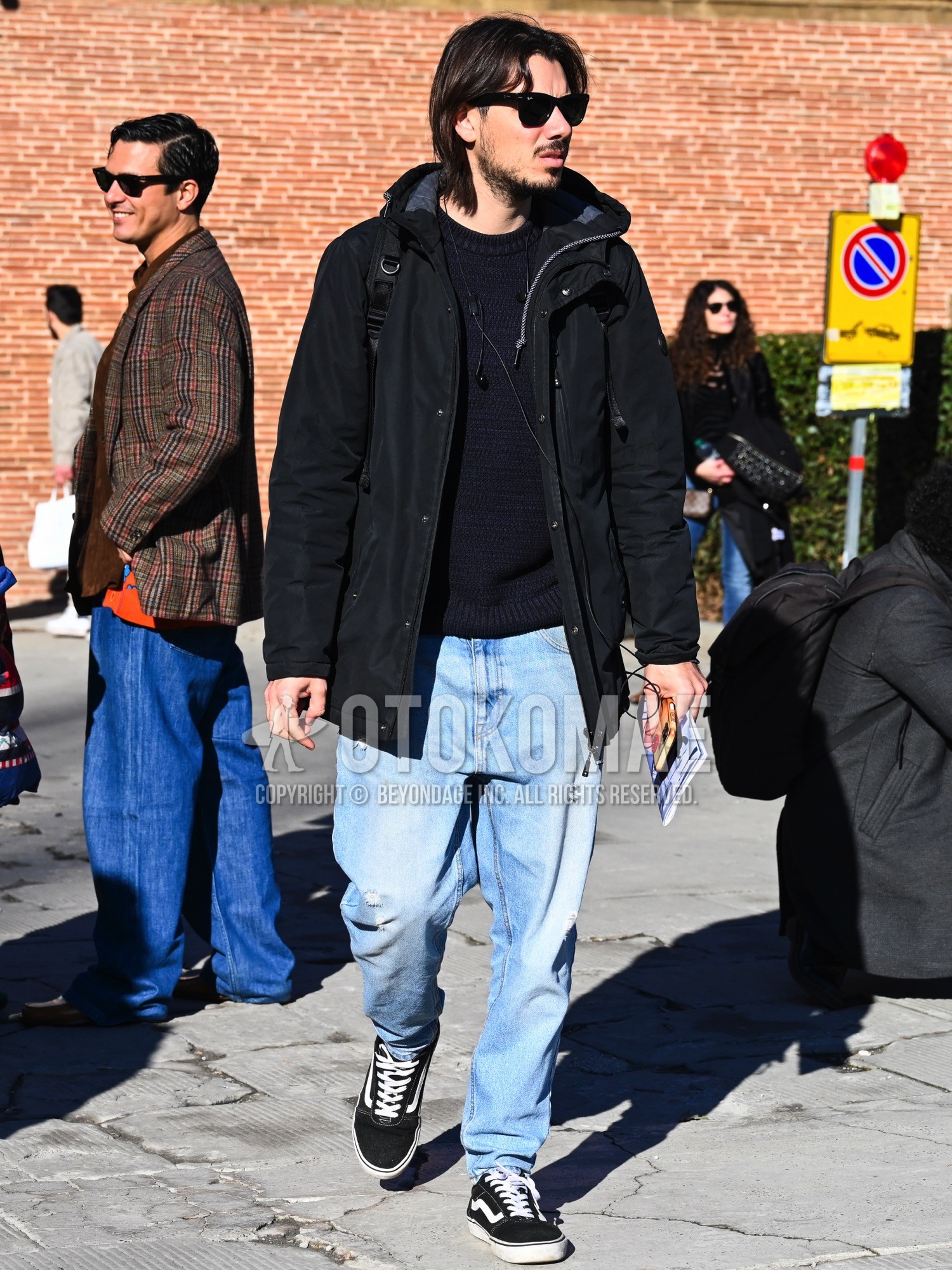 Men's autumn winter outfit with black plain sunglasses, black plain windbreaker, navy plain sweater, blue plain denim/jeans, blue plain damaged jeans, black low-cut sneakers.
