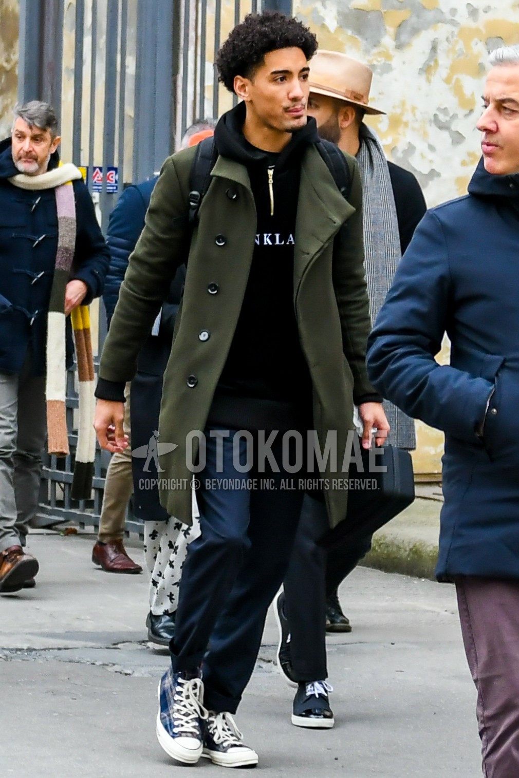 Men's autumn winter outfit with olive green plain stenkarrer coat, black graphic hoodie, navy plain slacks, navy high-cut sneakers.