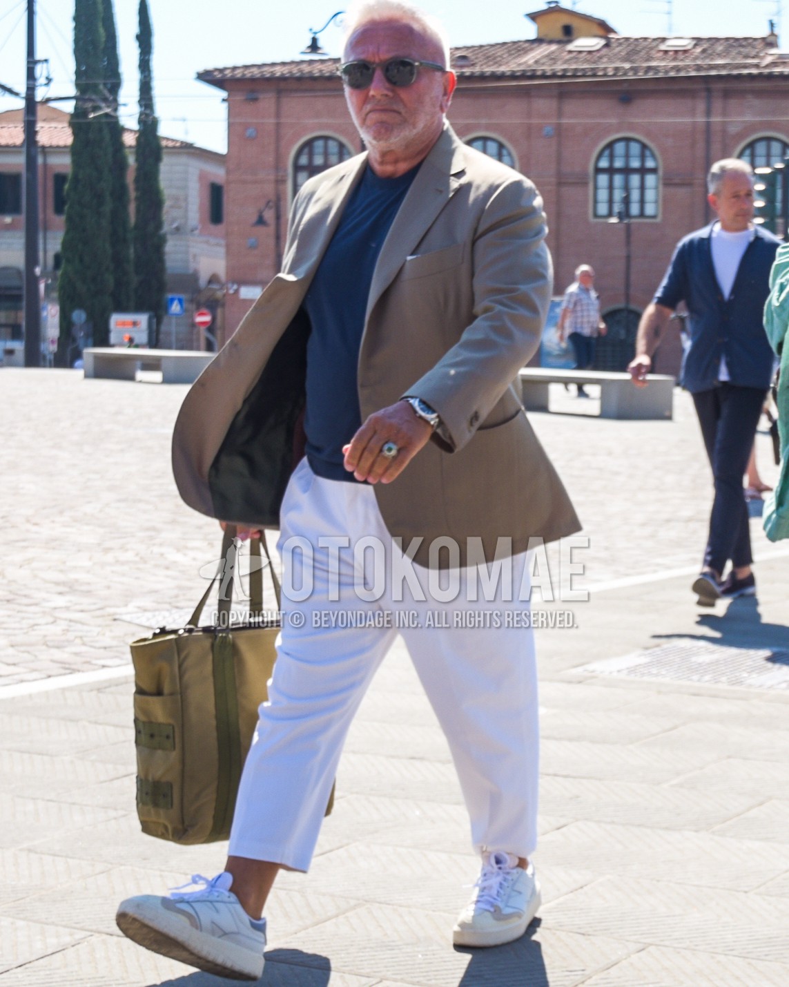Men's spring summer autumn outfit with gray plain sunglasses, beige plain tailored jacket, navy plain t-shirt, white plain slacks, white plain cropped pants, white low-cut sneakers, olive green plain tote bag.
