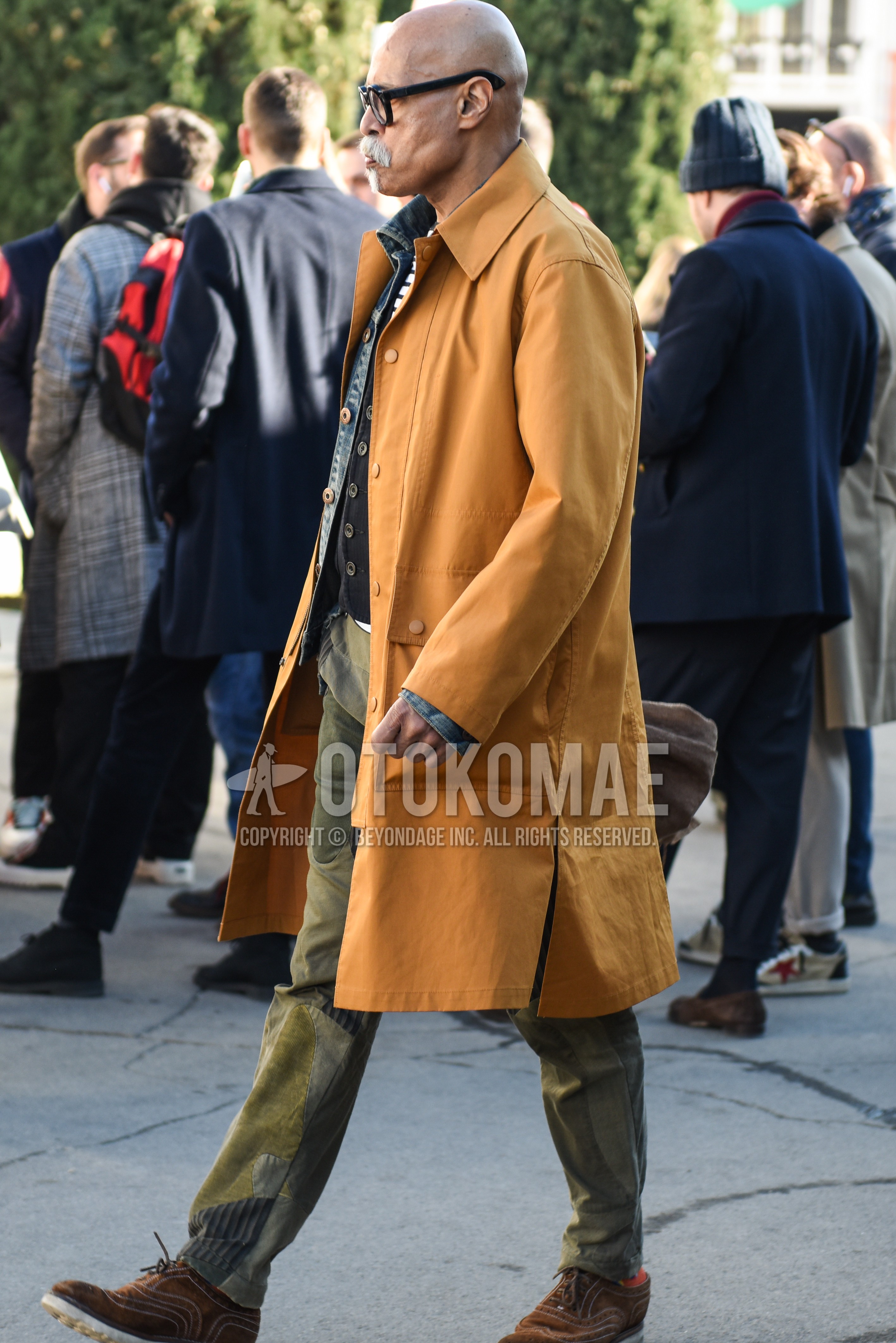 Men's autumn winter outfit with black plain glasses, yellow beige plain stenkarrer coat, blue plain denim shirt/chambray shirt, navy stripes gilet, white navy plain t-shirt, olive green bottoms chinos, brown wing-tip shoes leather shoes, brown brogue shoes leather shoes.