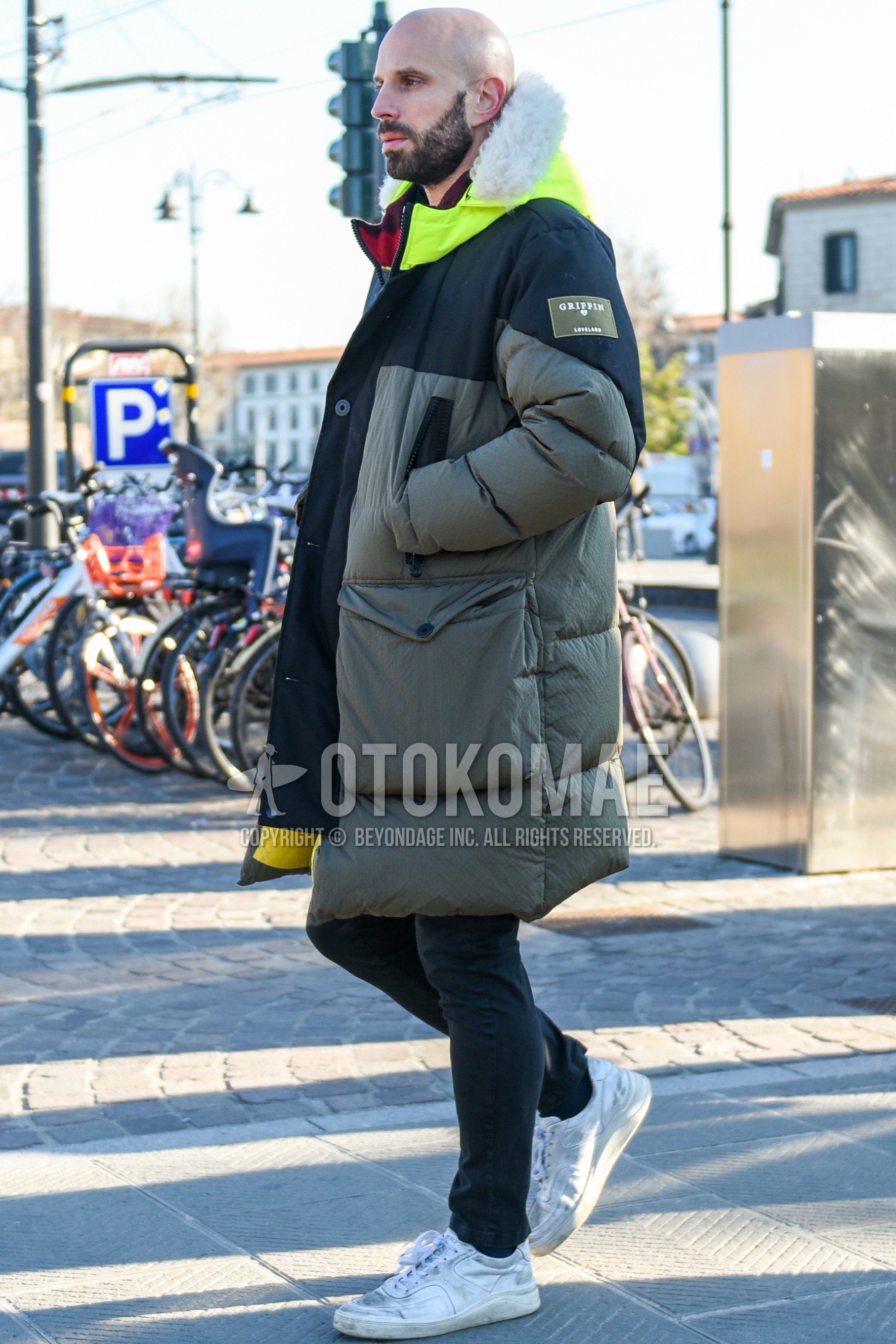 Men's winter outfit with olive green black plain down jacket, olive green black plain hooded coat, dark gray plain denim/jeans, white low-cut sneakers.
