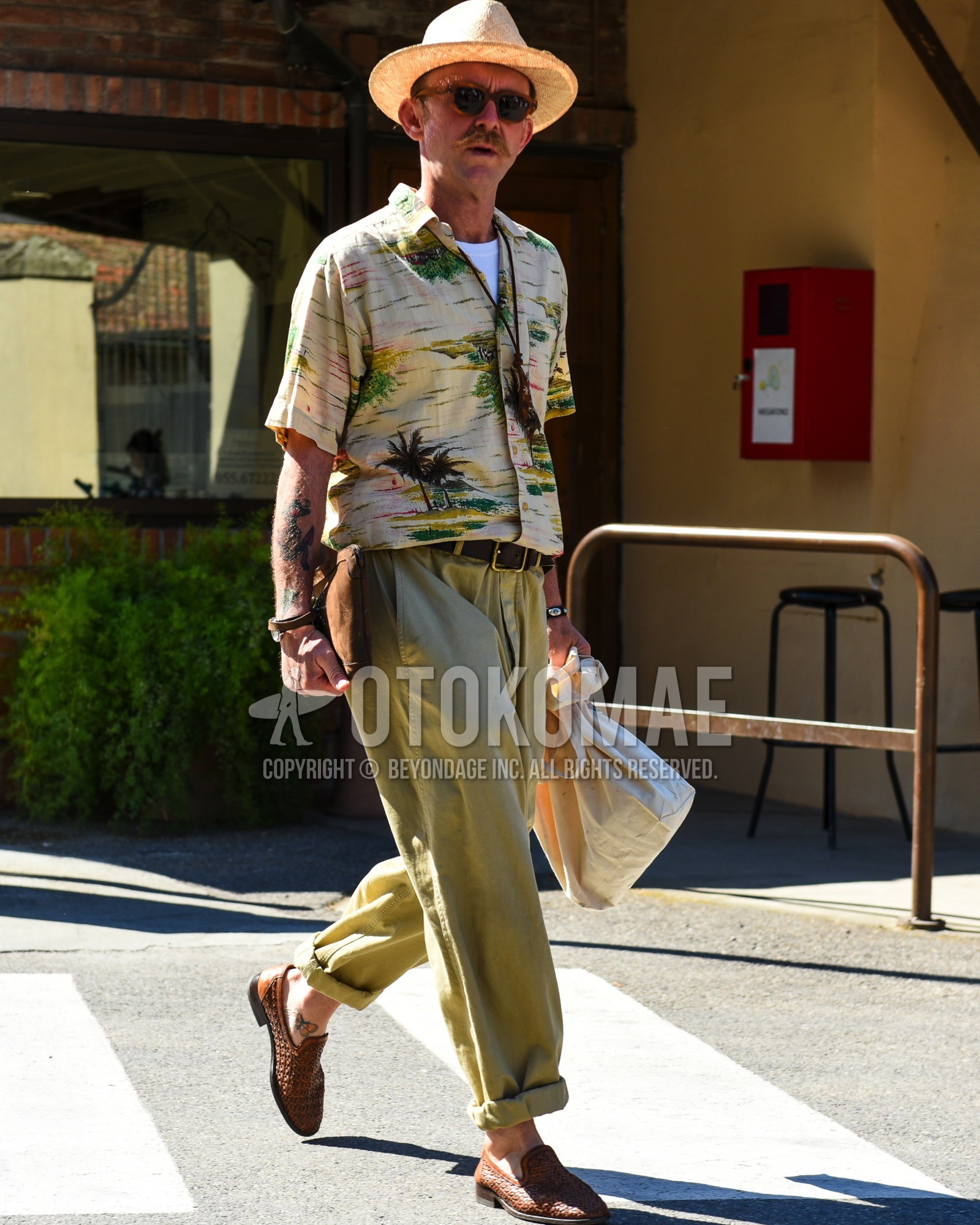 Men's spring summer outfit with beige plain hat, brown tortoiseshell sunglasses, beige graphic shirt, white plain t-shirt, brown plain leather belt, olive green plain chinos, brown  loafers leather shoes, white plain tote bag, brown plain clutch bag/second bag/drawstring bag.