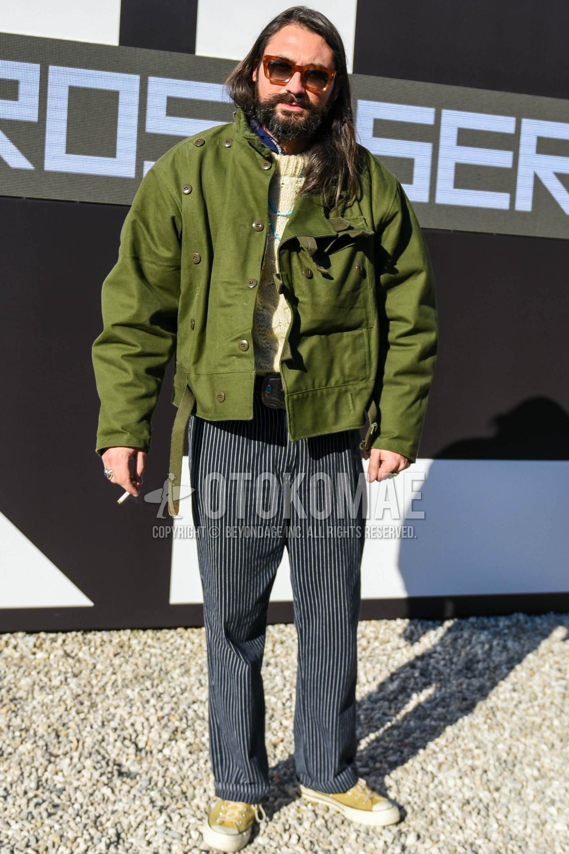 Men's autumn winter outfit with brown tortoiseshell sunglasses, olive green plain field jacket/hunting jacket, white plain sweater, navy stripes slacks, yellow low-cut sneakers.