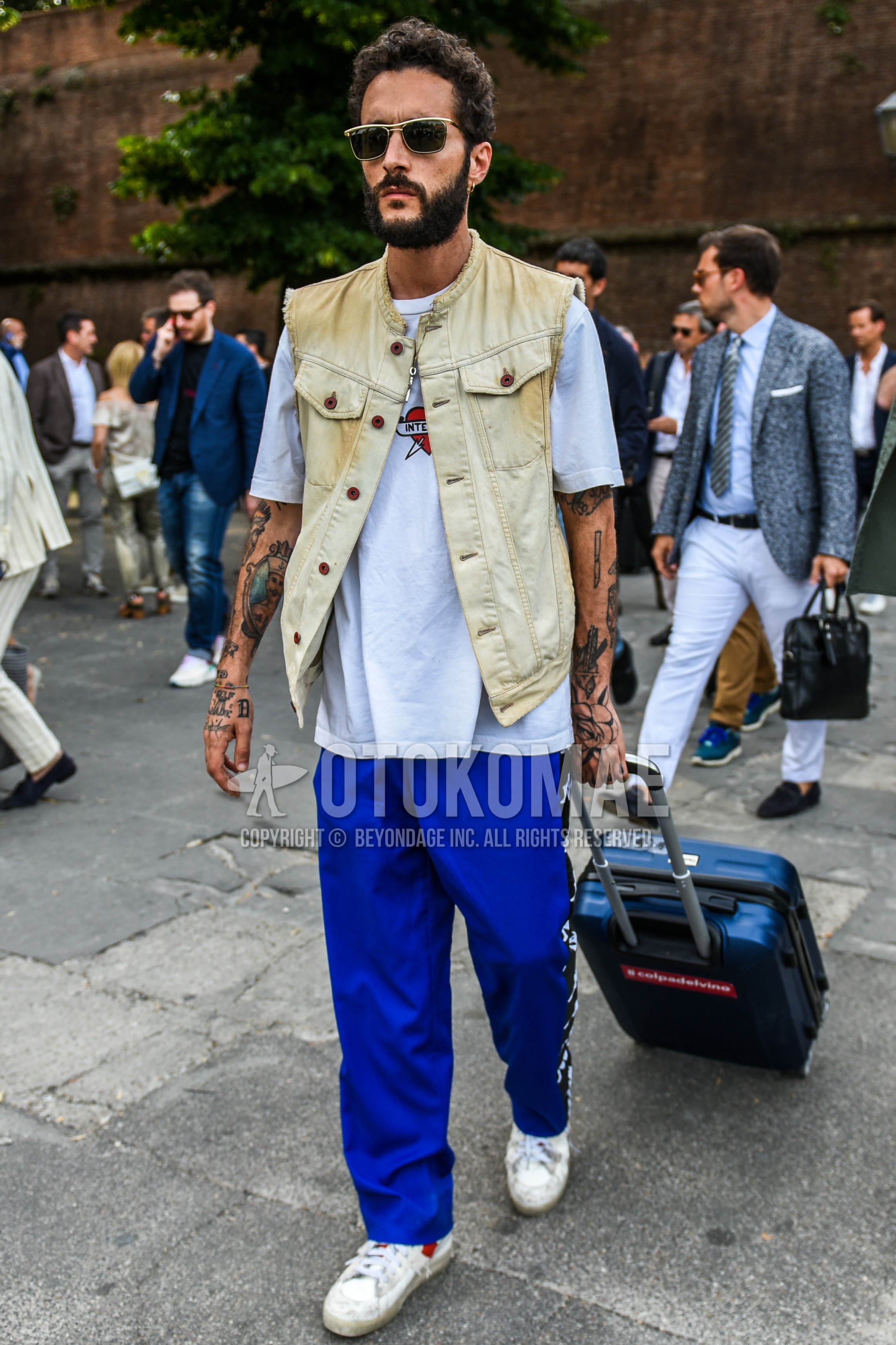 Men's summer outfit with gold plain sunglasses, beige plain denim jacket, white graphic t-shirt, blue bottoms sideline pants, white low-cut sneakers, navy plain suitcase.