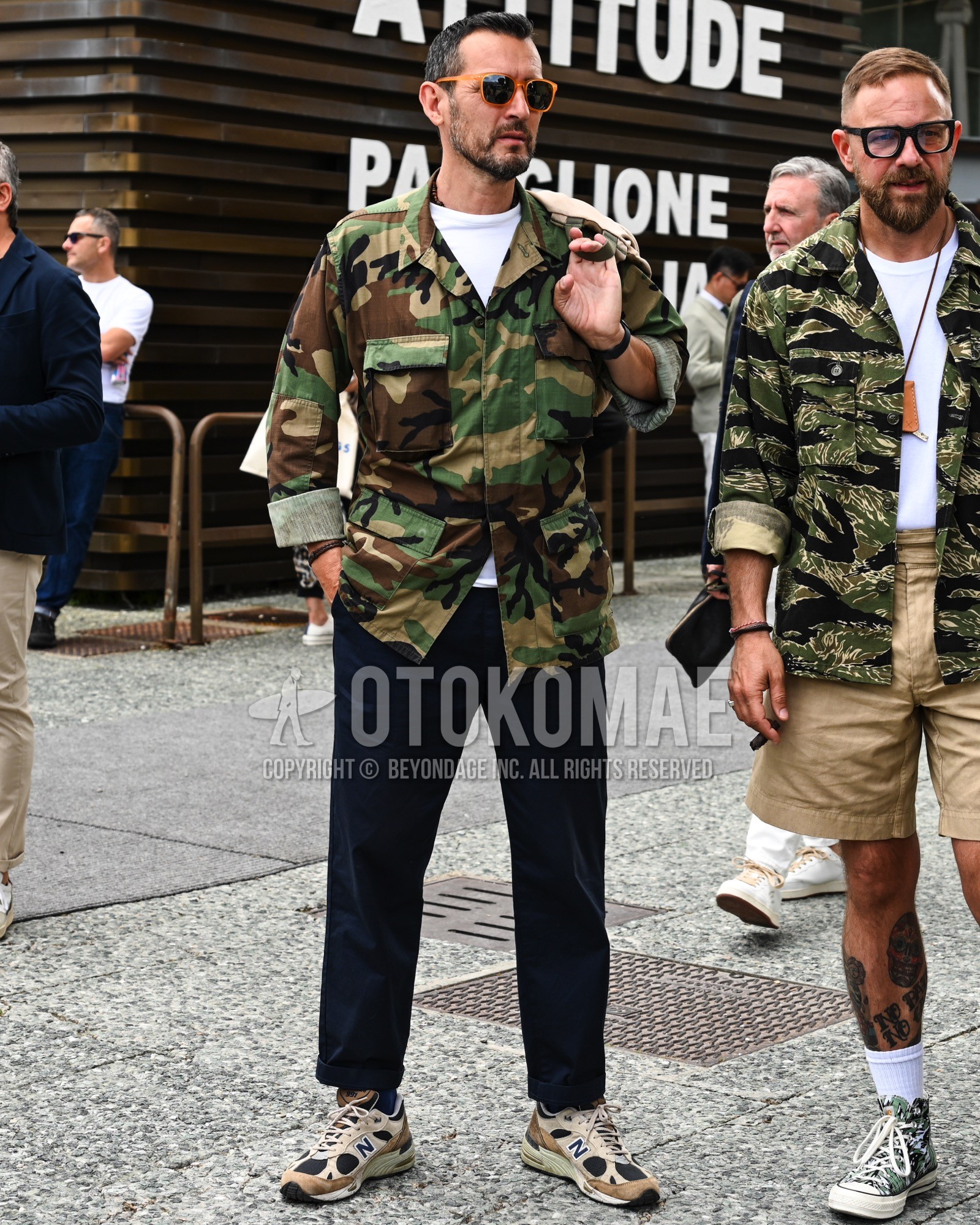 Men's spring summer autumn outfit with black plain sunglasses, olive green camouflage military jacket, white plain t-shirt, black plain chinos, beige low-cut sneakers, beige plain tote bag.
