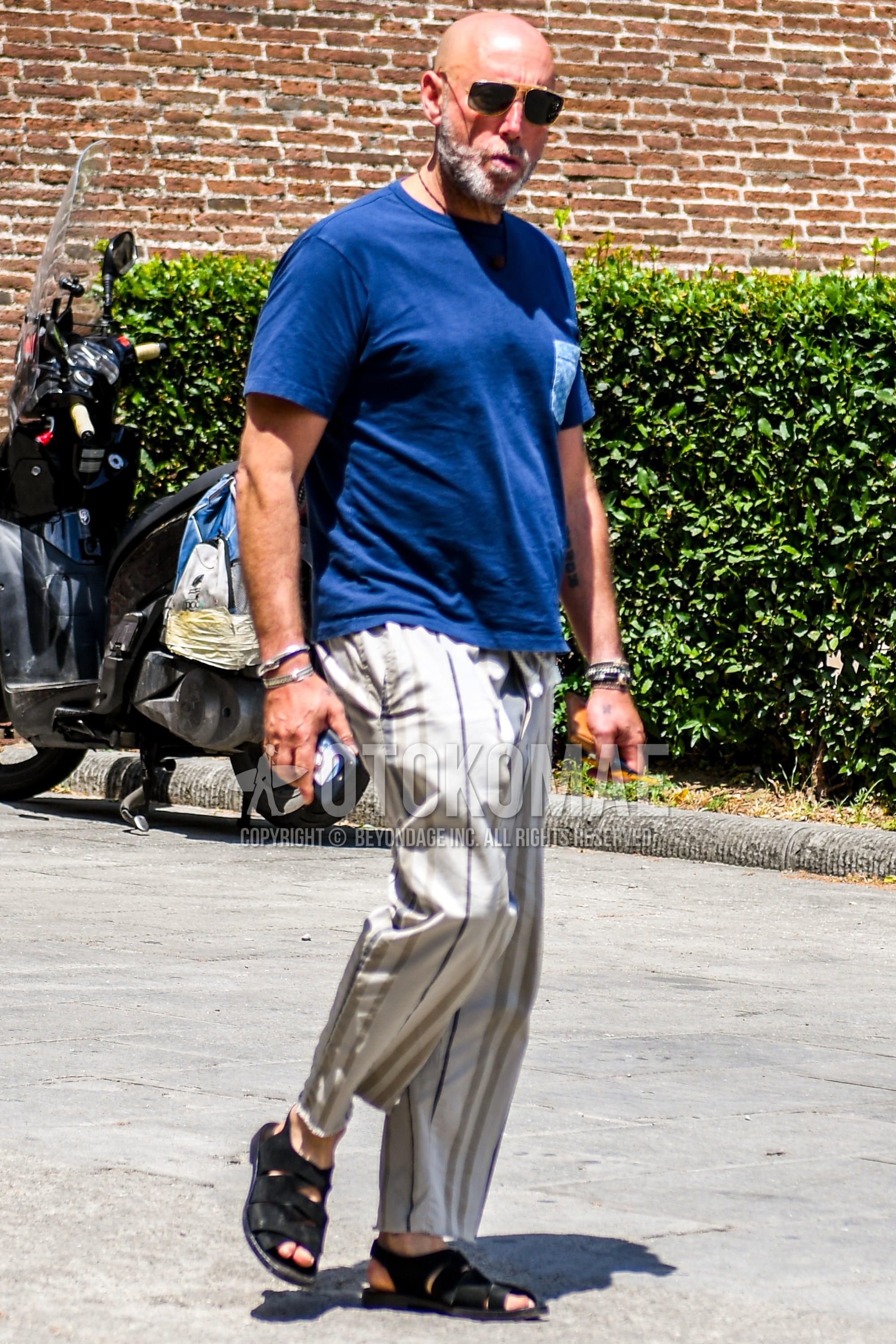 Men's summer outfit with plain sunglasses, blue plain t-shirt, beige multi-color stripes wide pants, black leather sandals.