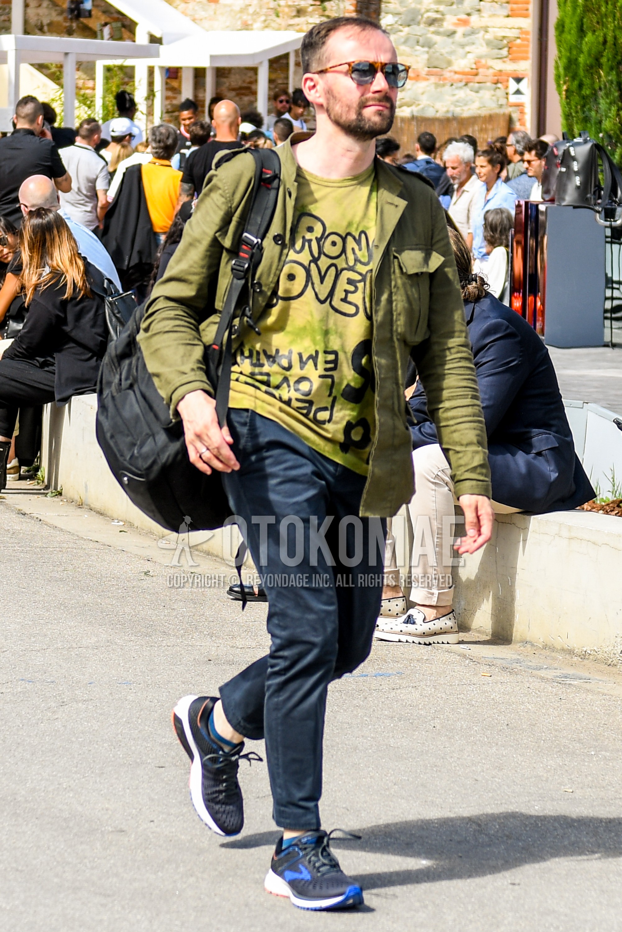 Men's spring summer autumn outfit with olive green plain M-65, olive green graphic t-shirt, navy plain chinos, navy plain cotton pants, black low-cut sneakers, black plain backpack.