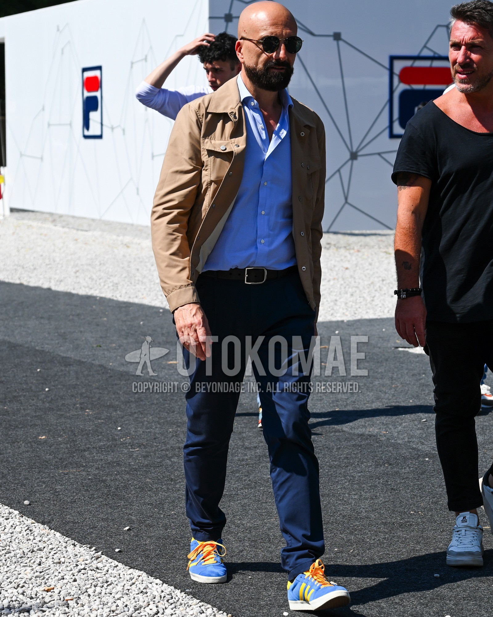 Men's spring summer autumn outfit with black plain sunglasses, beige plain field jacket/hunting jacket, light blue plain shirt, black plain leather belt, navy plain chinos, light blue low-cut sneakers.