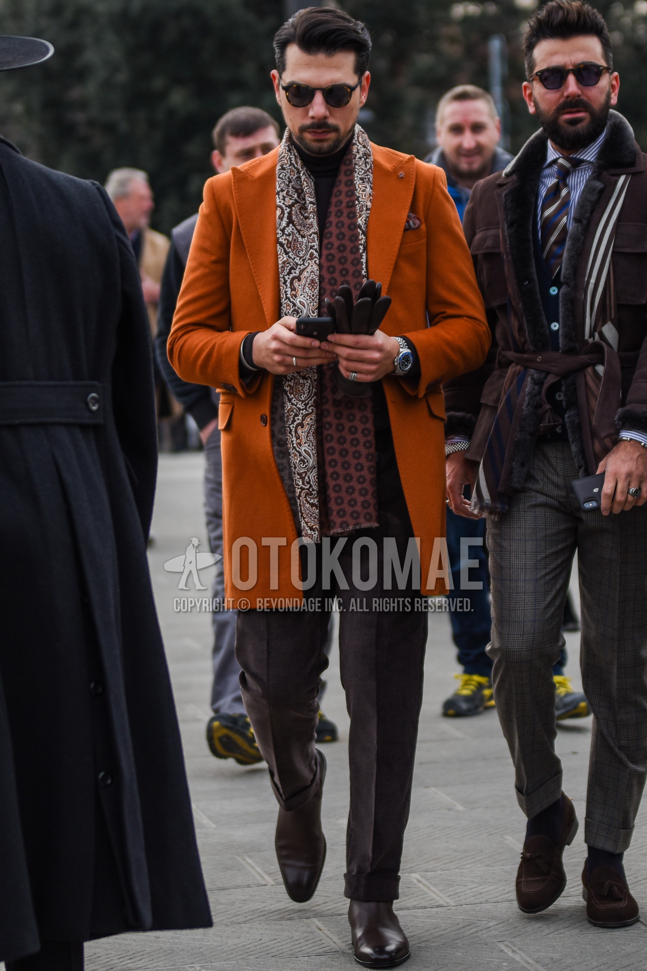 Men's autumn winter outfit with brown tortoiseshell sunglasses, brown paisley scarf, brown small crest scarf, orange plain chester coat, brown plain turtleneck knit, brown plain slacks, brown side-gore boots.