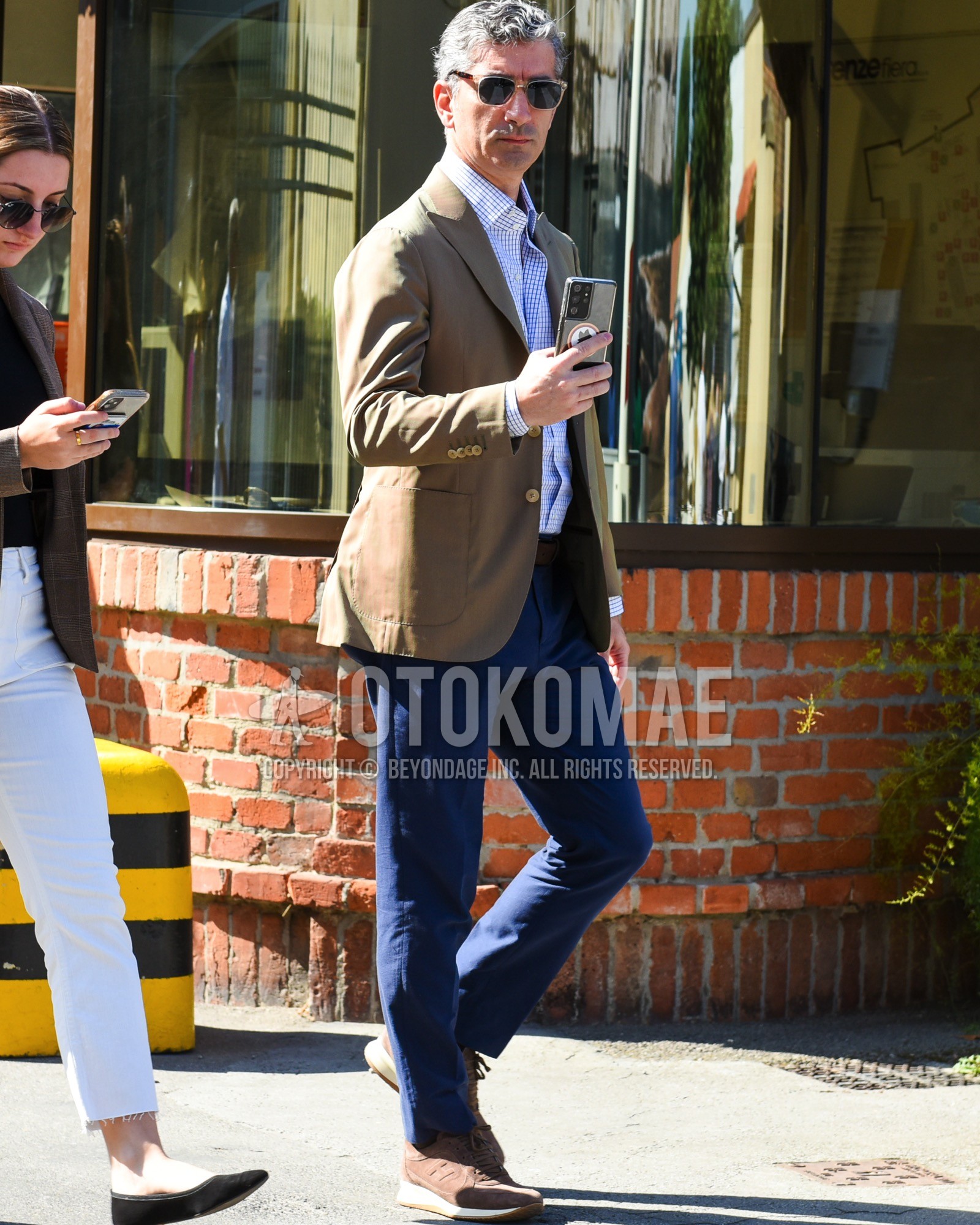 Men's spring summer outfit with clear brown tortoiseshell sunglasses, brown plain tailored jacket, blue white check shirt, brown plain leather belt, navy plain slacks, brown low-cut sneakers.