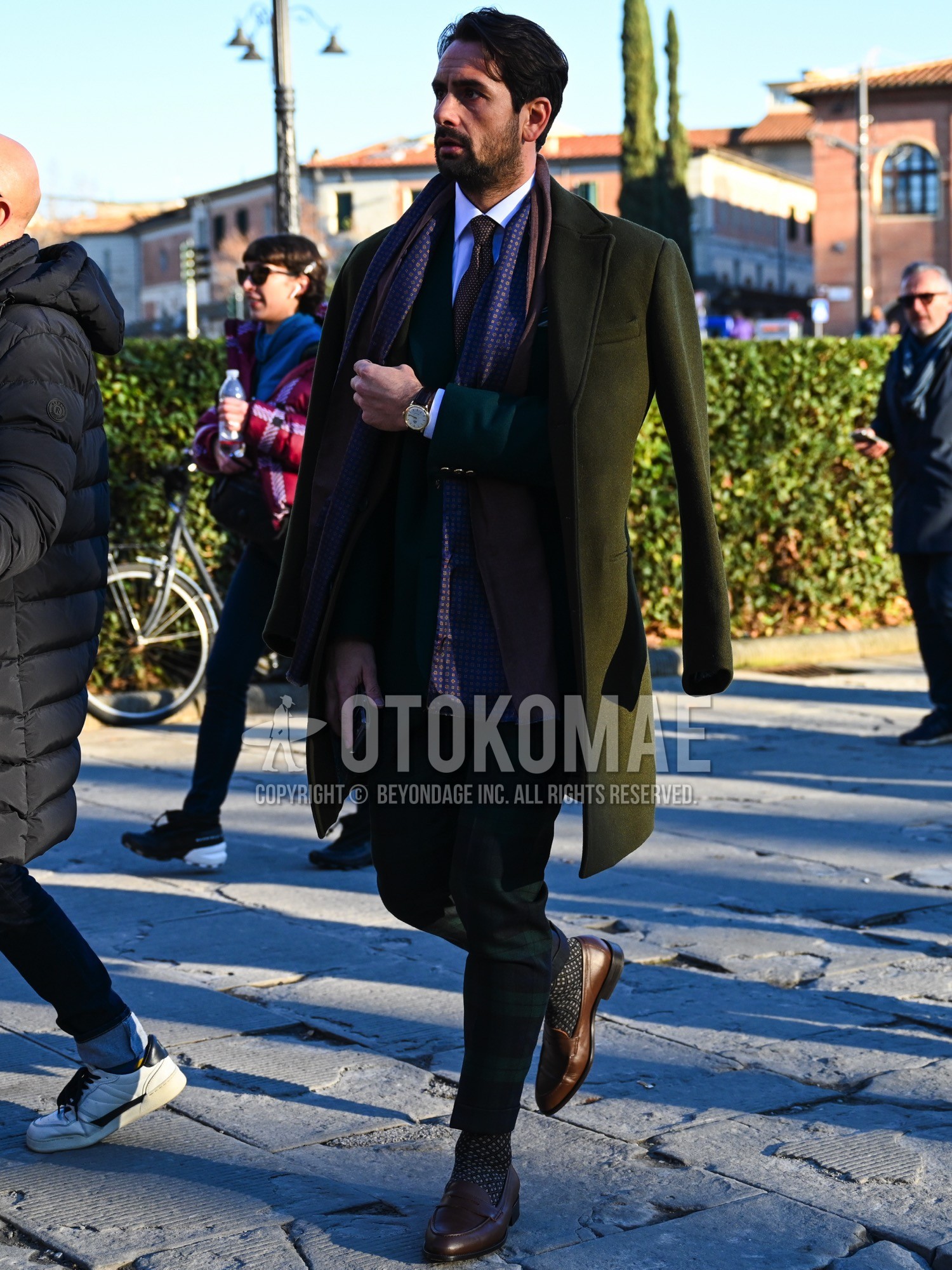 Men's autumn winter outfit with purple dots scarf, olive green plain chester coat, white plain shirt, olive green plain tailored jacket, green check ankle pants, black dots socks, brown coin loafers leather shoes, brown small crest necktie.