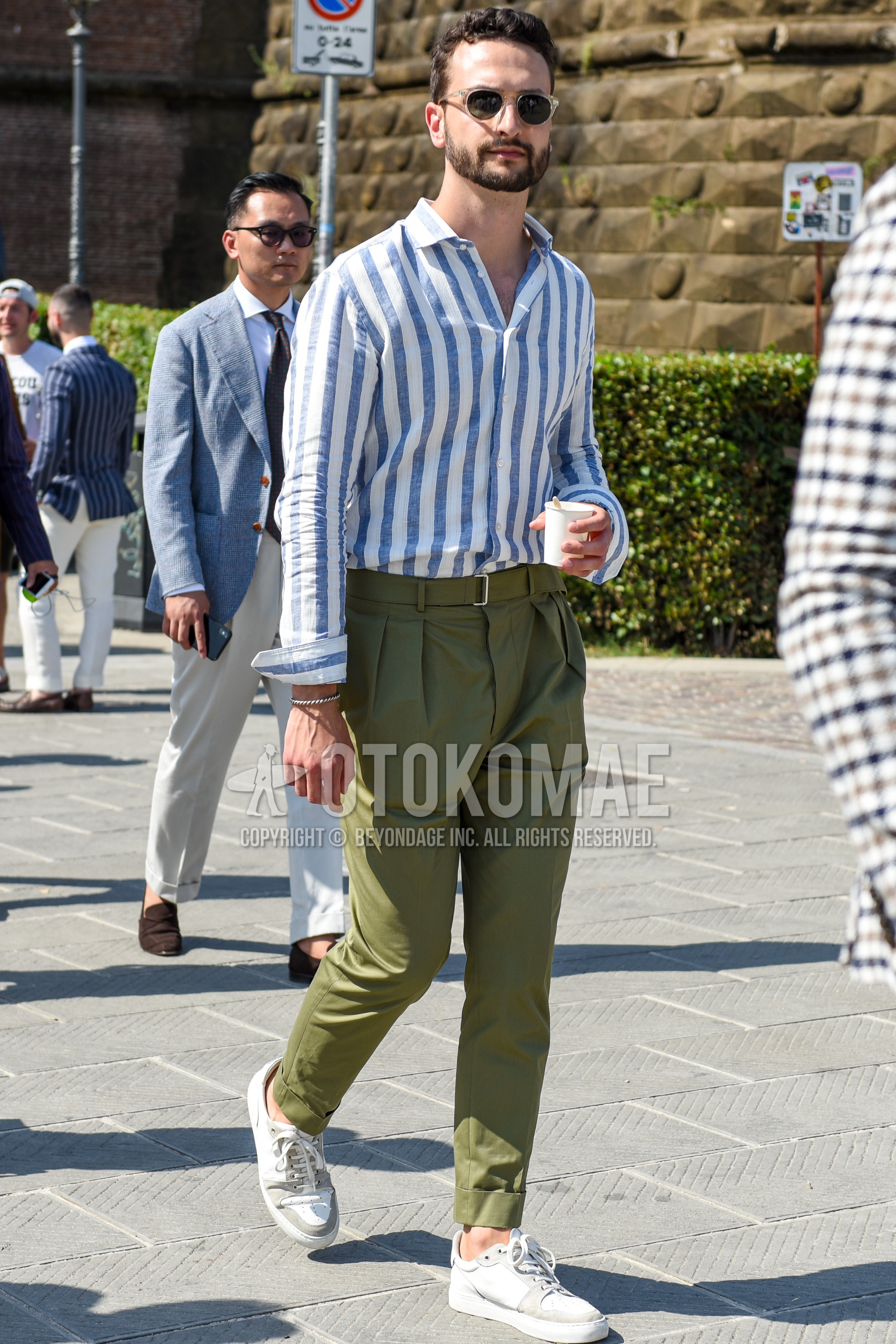 Men's spring summer autumn outfit with beige plain sunglasses, blue white stripes shirt, olive green plain beltless pants, white low-cut sneakers.