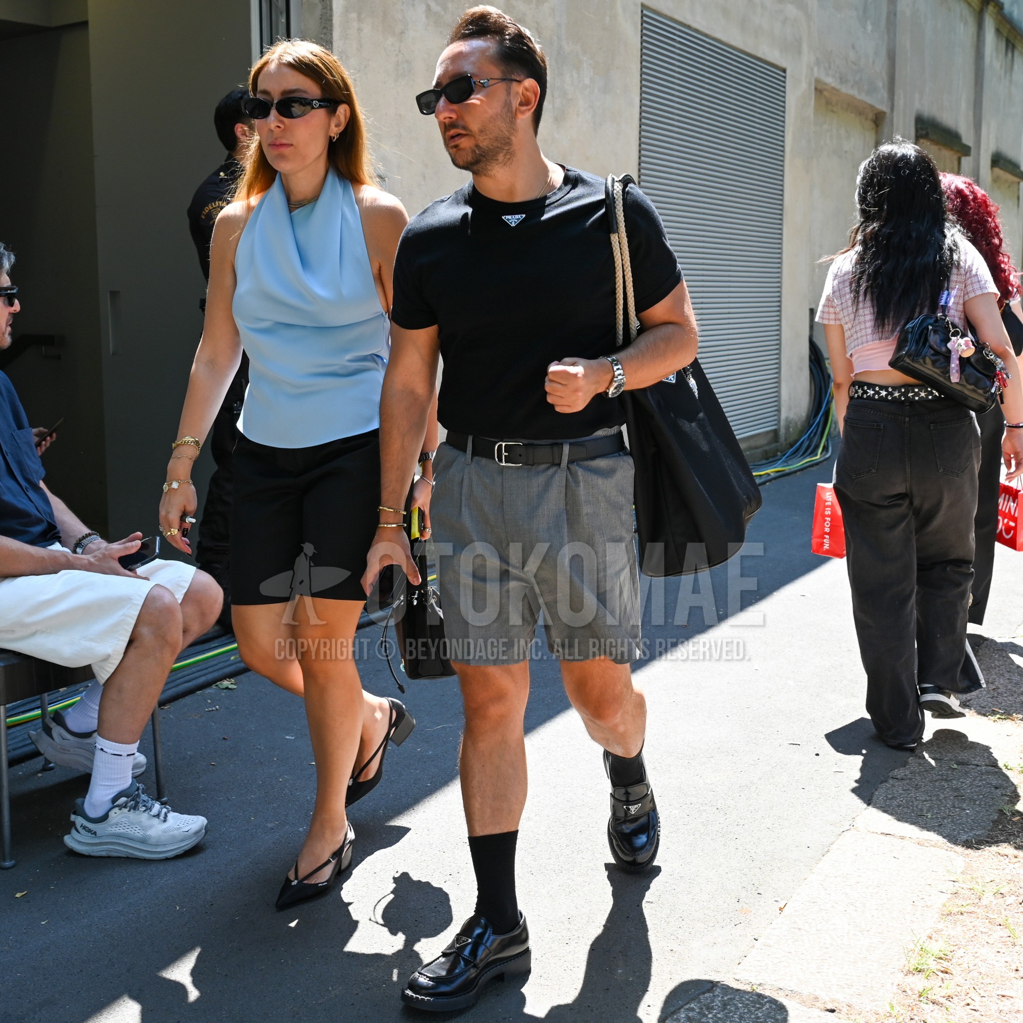 プラダの黒無地のサングラス、プラダの黒デカロゴのTシャツ、黒無地のレザーベルト、グレーチェックのショートパンツ、グレーチェックのスラックス、黒無地のソックス、プラダの黒ローファーの革靴、プラダの黒無地のトートバッグを合わせた夏のメンズコーデ・着こなし。