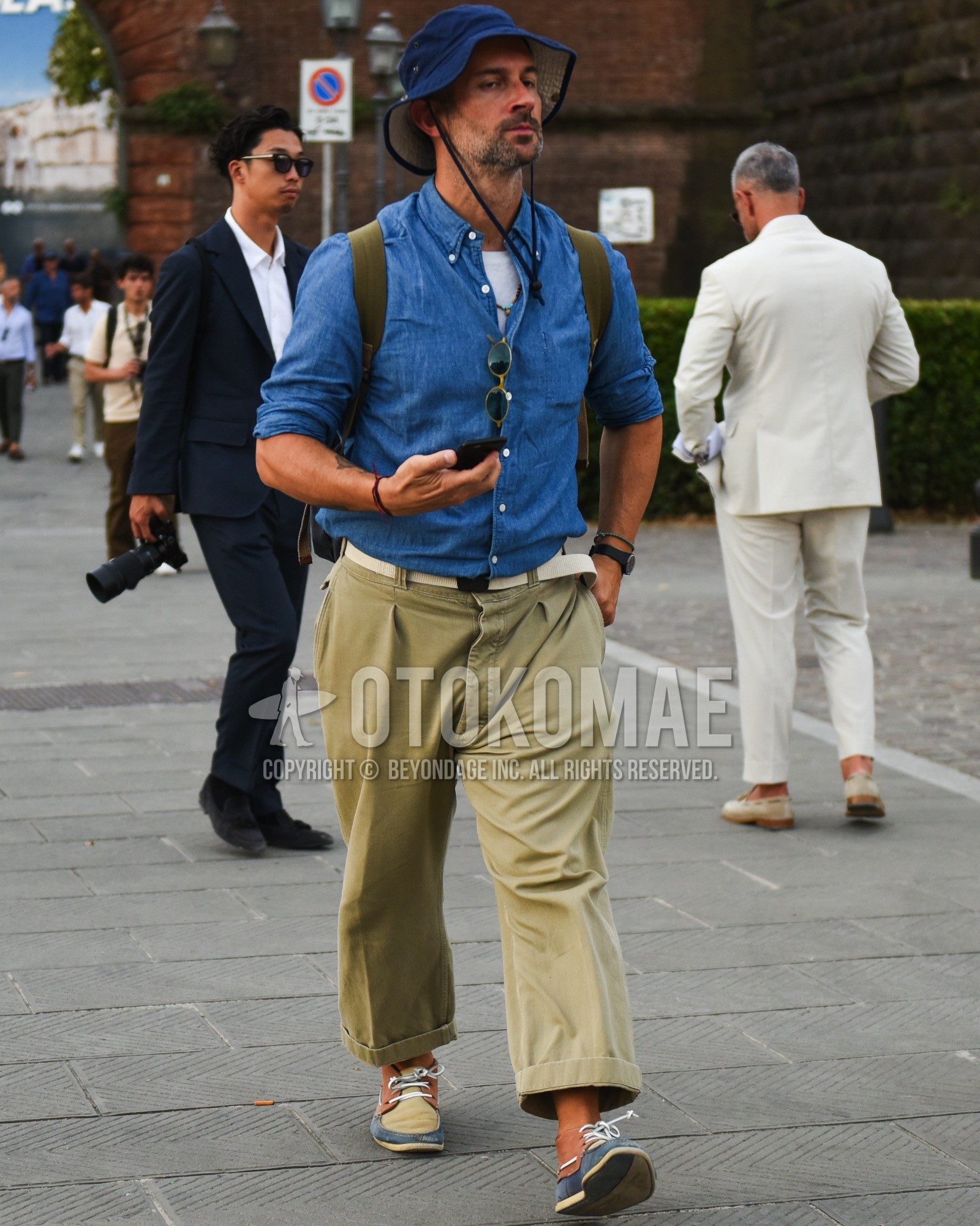 Men's spring summer outfit with blue plain bucket hat, blue plain denim shirt/chambray shirt, white plain t-shirt, white plain tape belt, beige plain cotton pants, beige plain cropped pants, beige plain wide pants, beige brown low-cut sneakers, olive green plain backpack.