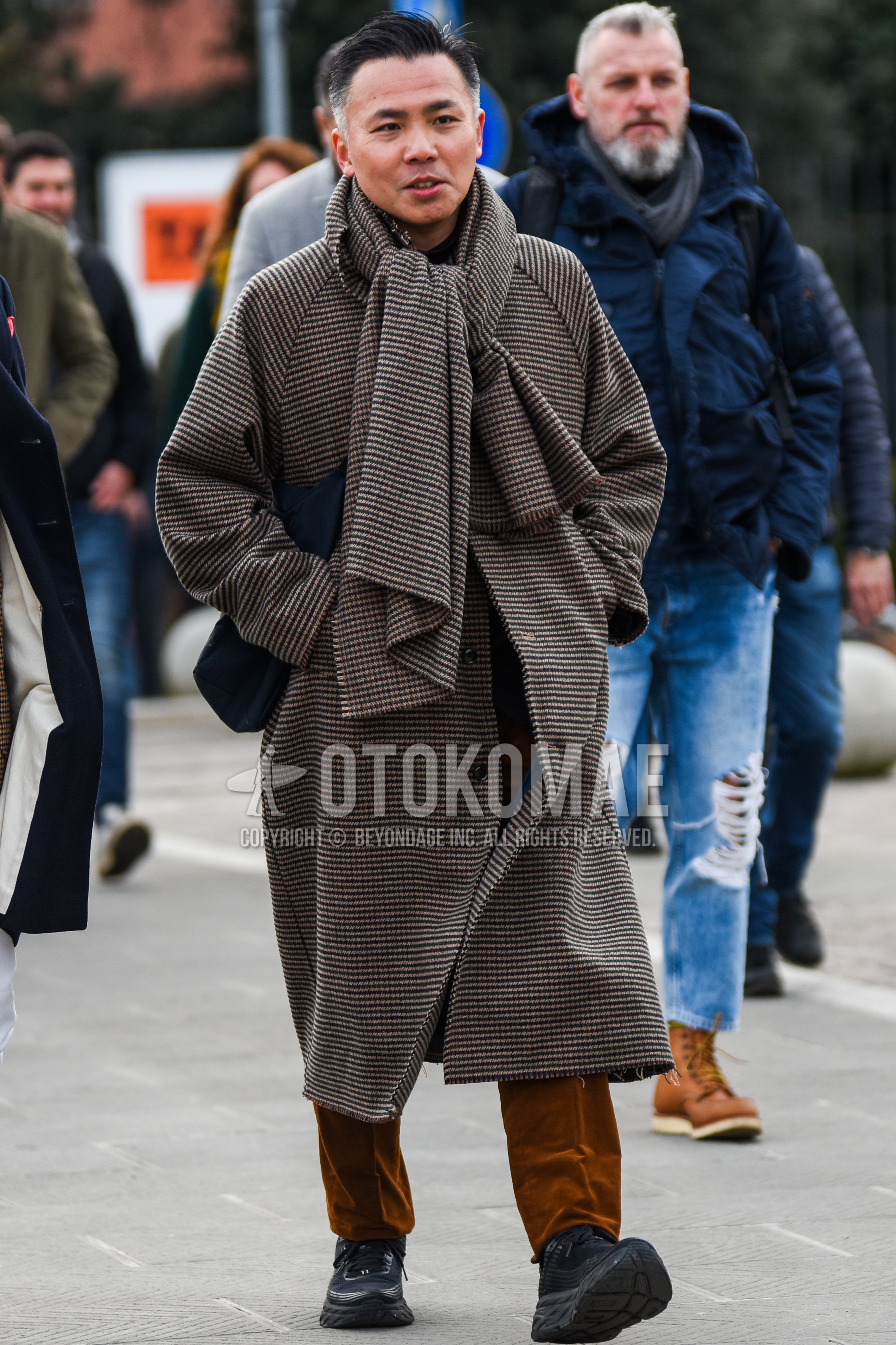 Men's winter outfit with brown check scarf, brown plain trench coat, brown plain chinos, black low-cut sneakers, black plain shoulder bag.