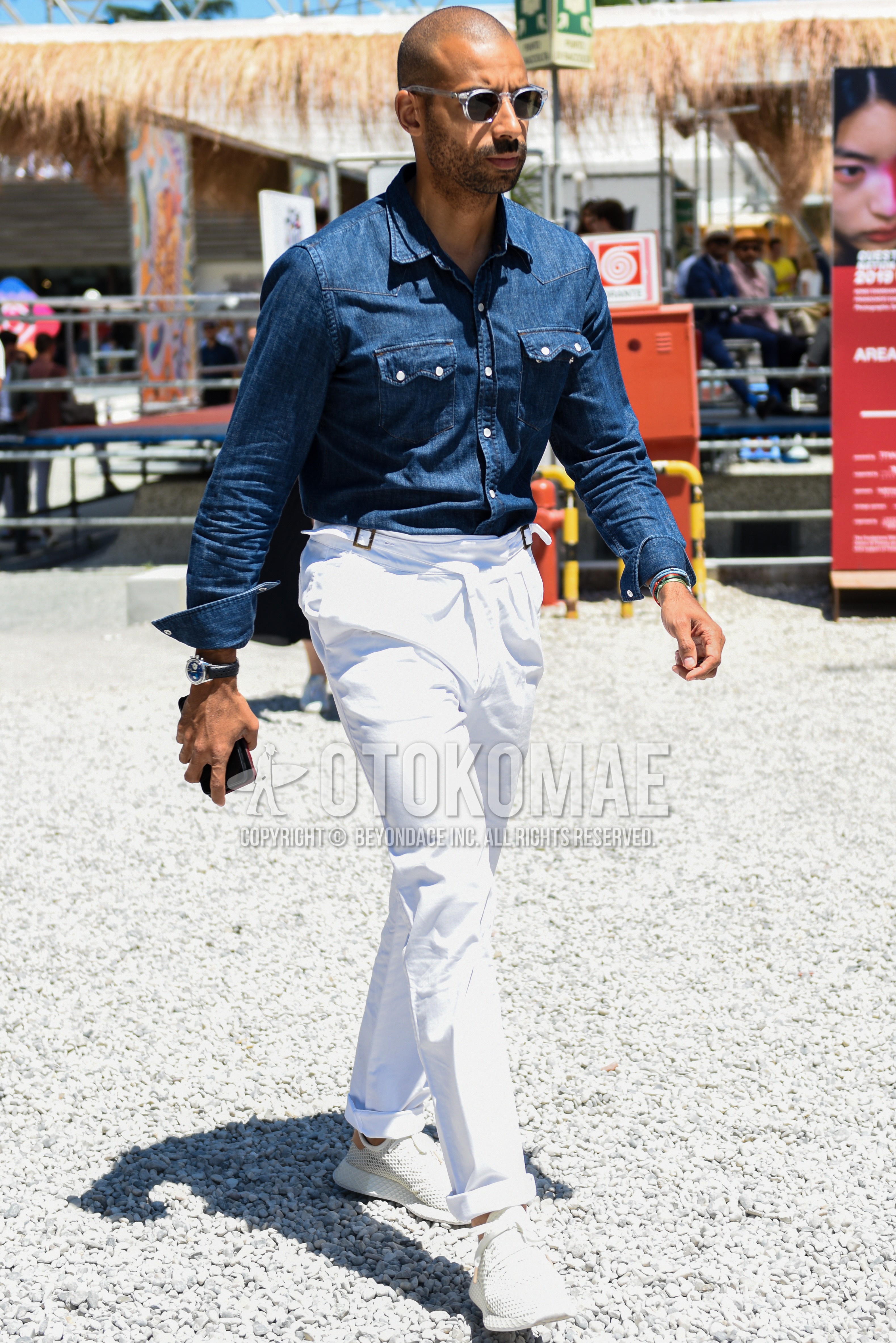 Men's spring summer outfit with clear plain sunglasses, blue plain denim shirt/chambray shirt, white plain beltless pants, white low-cut sneakers.