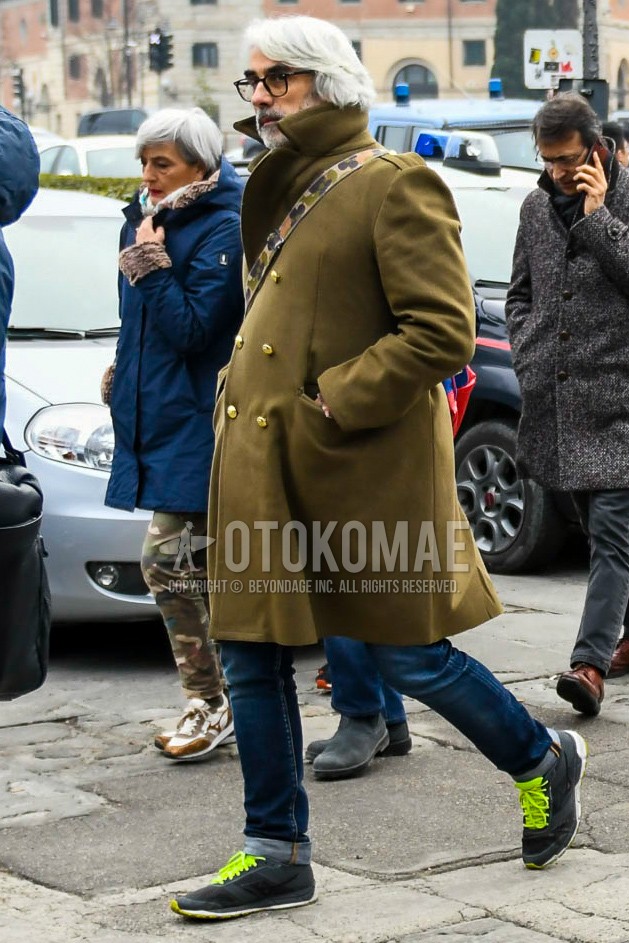 Men's winter outfit with black plain glasses, olive green plain ulster coat, navy plain denim/jeans, gray low-cut sneakers.