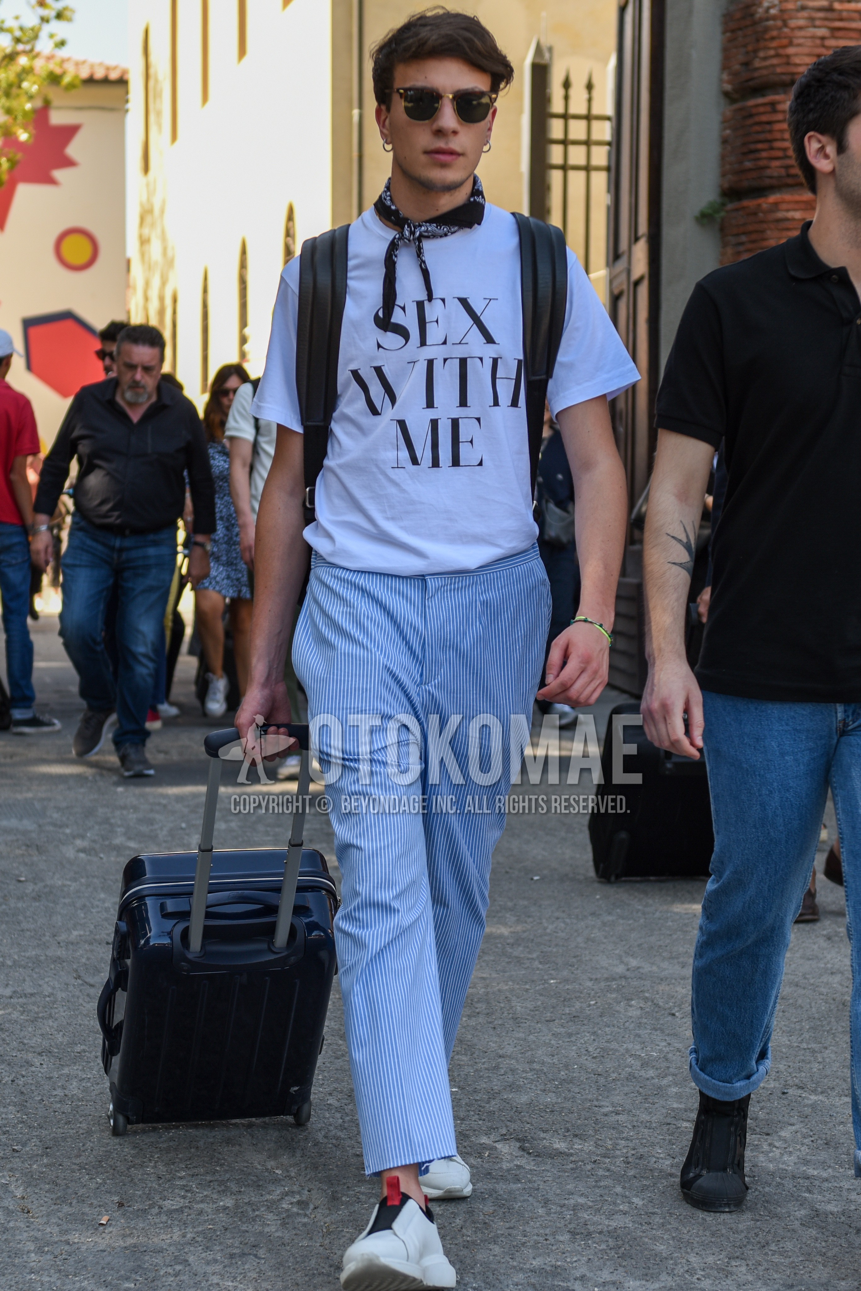Men's summer outfit with black plain sunglasses, black paisley scarf, white graphic t-shirt, light blue stripes ankle pants, white slip-on sneakers.