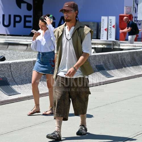 ブラウンレタードのベースボールキャップ、ブラウンべっ甲のサングラス、グリーン無地のカジュアルベスト、白無地のV ネック、白無地のTシャツ、ブラウンレオパードのショートパンツ、ベージュ無地のソックス、グリーンサンダルを合わせた夏のメンズコーデ・着こなし。