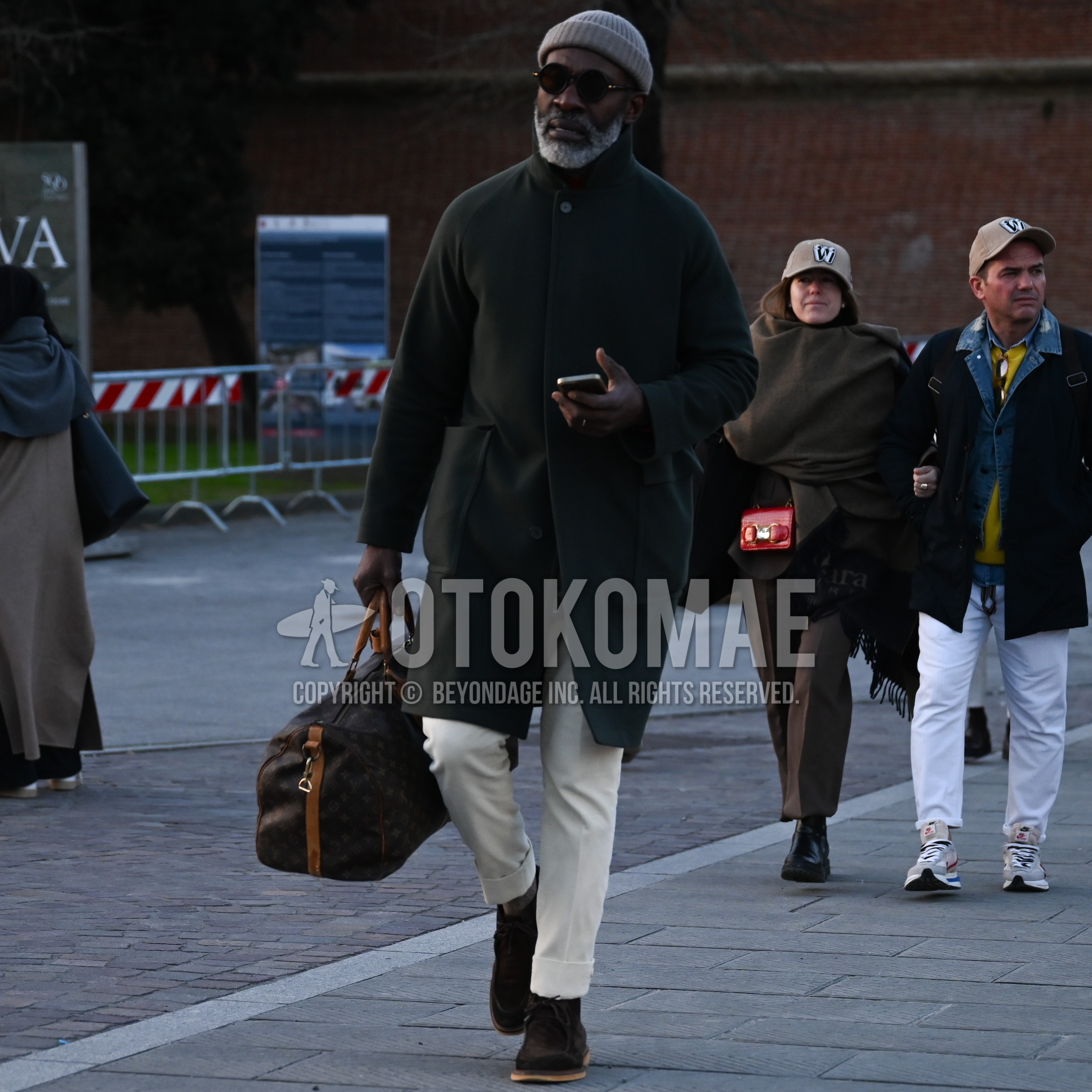 Men's autumn winter outfit with gray plain knit cap, black plain sunglasses, black plain stand collar coat, white beige plain slacks, brown  boots, brown whole pattern boston bag.