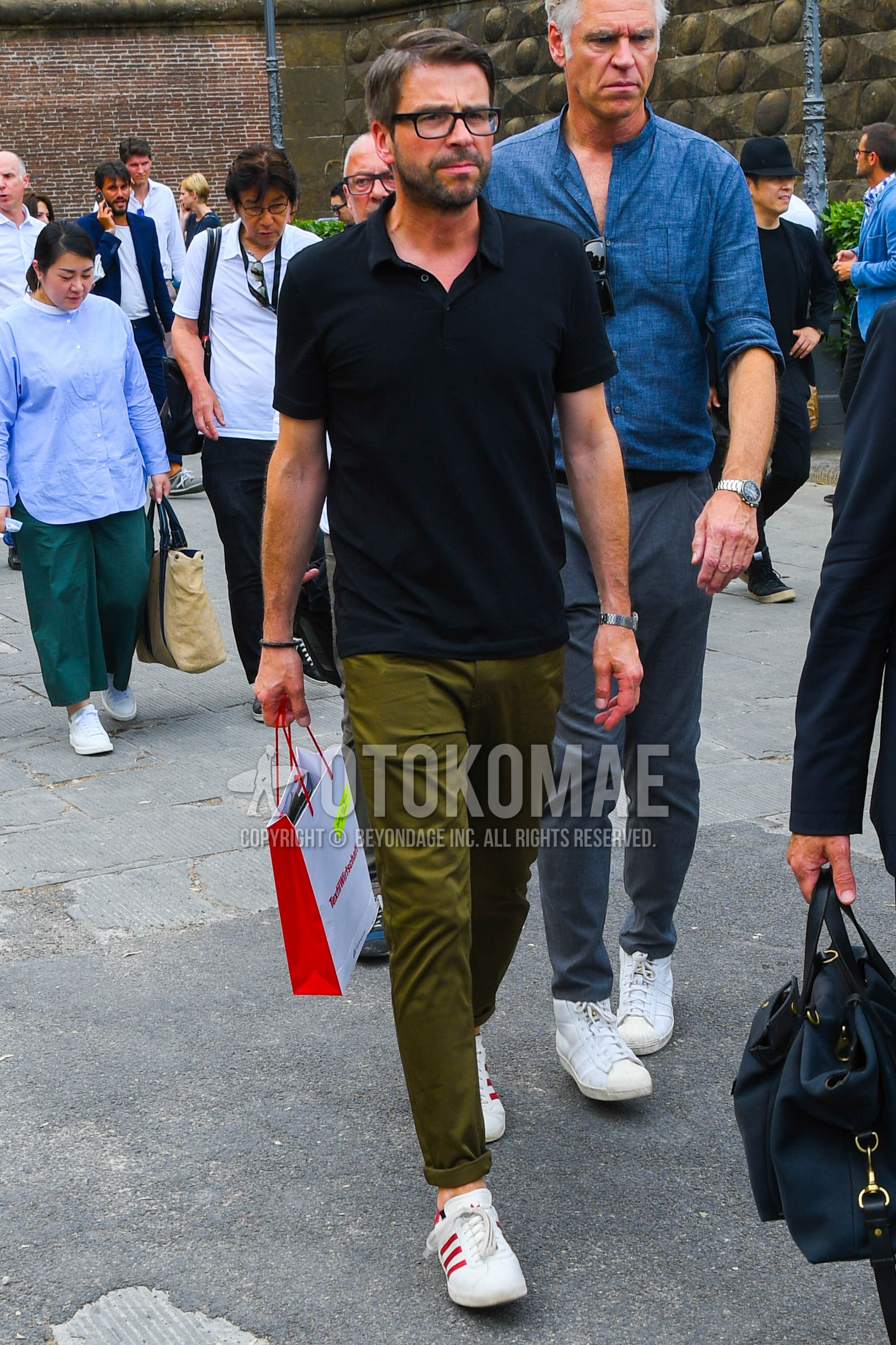 Men's spring summer outfit with black plain glasses, black plain polo shirt, olive green plain chinos, white low-cut sneakers.