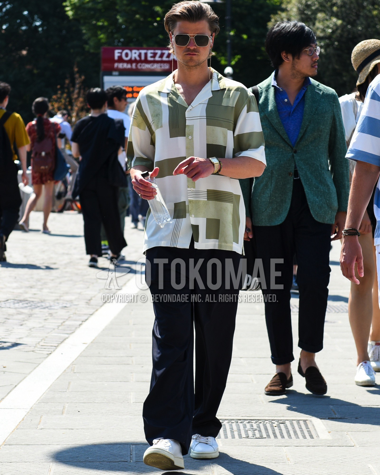 Men's spring summer outfit with gold plain sunglasses, olive green white graphic shirt, black plain slacks, white low-cut sneakers.