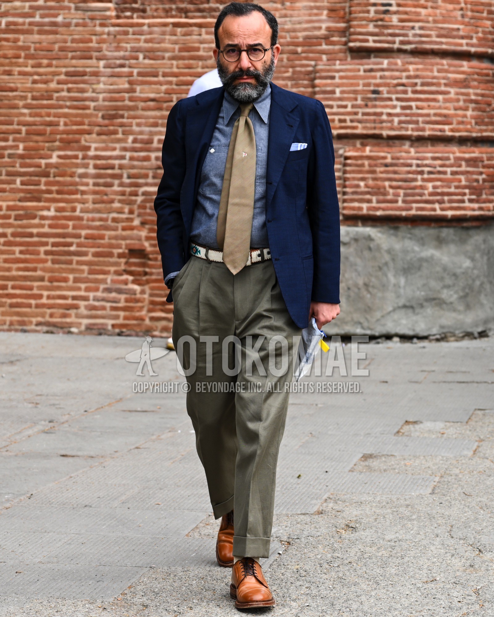 Men's spring summer autumn outfit with clear plain sunglasses, navy plain tailored jacket, light blue plain denim shirt/chambray shirt, white belt leather belt, olive green plain chinos, olive green plain pleated pants, brown plain toe leather shoes.