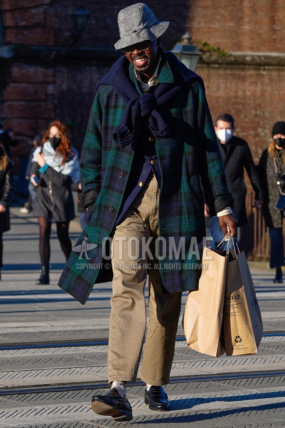Men's spring winter outfit with gray check cap, clear plain glasses, green check stenkarrer coat, navy plain tailored jacket, yellow plain shirt, beige plain chinos, beige socks socks, black coin loafers leather shoes.