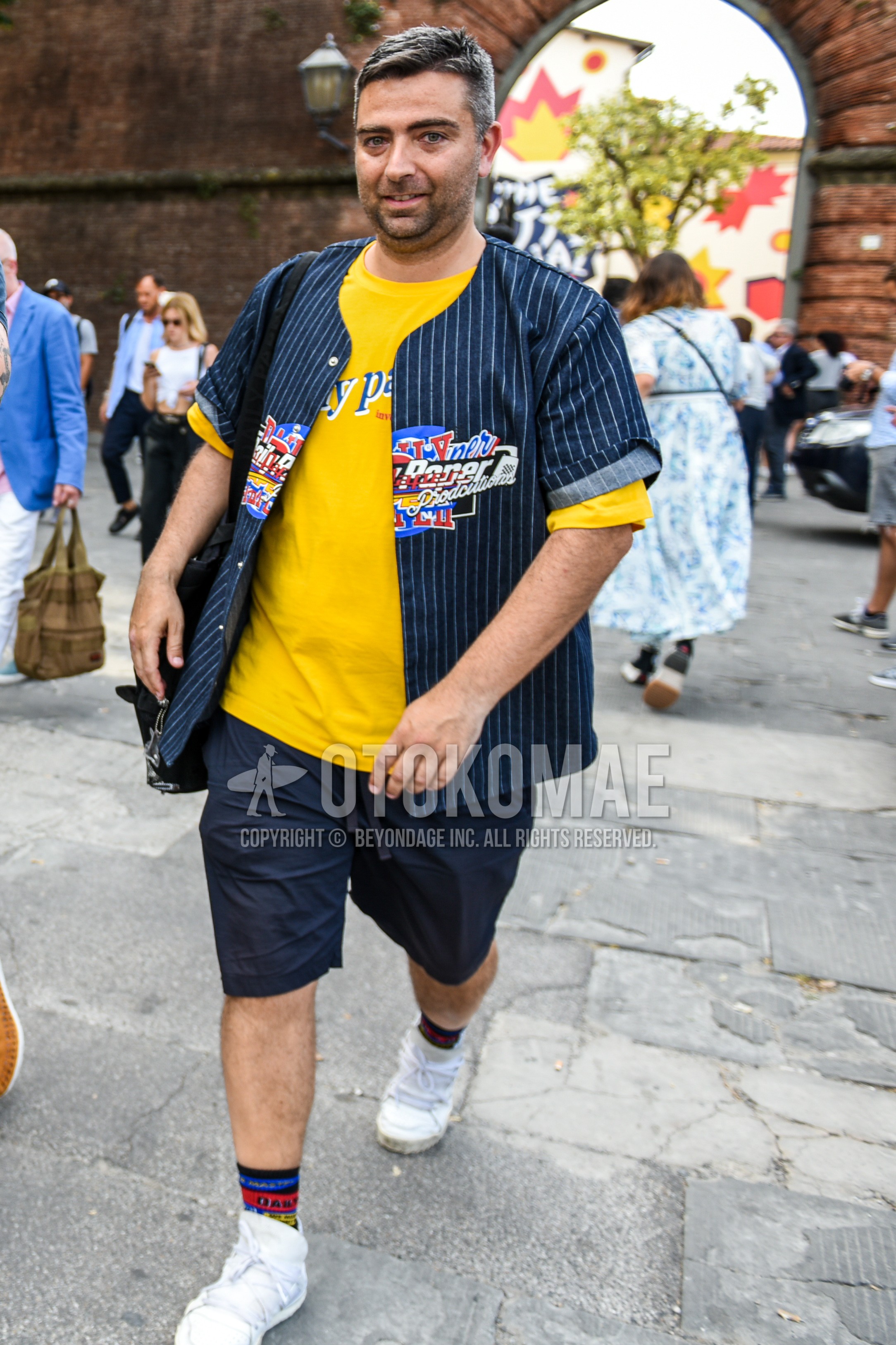 Men's summer outfit with yellow graphic t-shirt, navy stripes shirt, black plain short pants, multi-color plain socks, white low-cut sneakers.