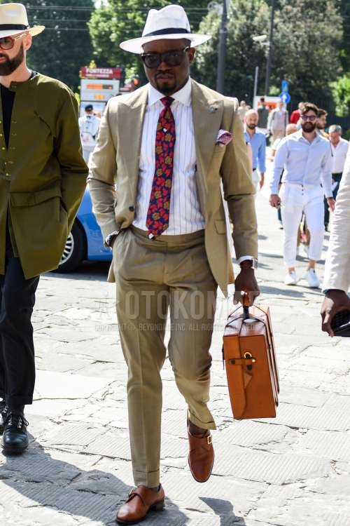 白無地のハット、黒無地のサングラス、白ストライプのシャツ、ブラウンモンクシューズの革靴、ブラウン無地のブリーフケース/ハンドバッグ、ベージュ無地のスーツ、レッド・ネイビーネクタイのネクタイを合わせた春夏秋のメンズコーデ・着こなし。