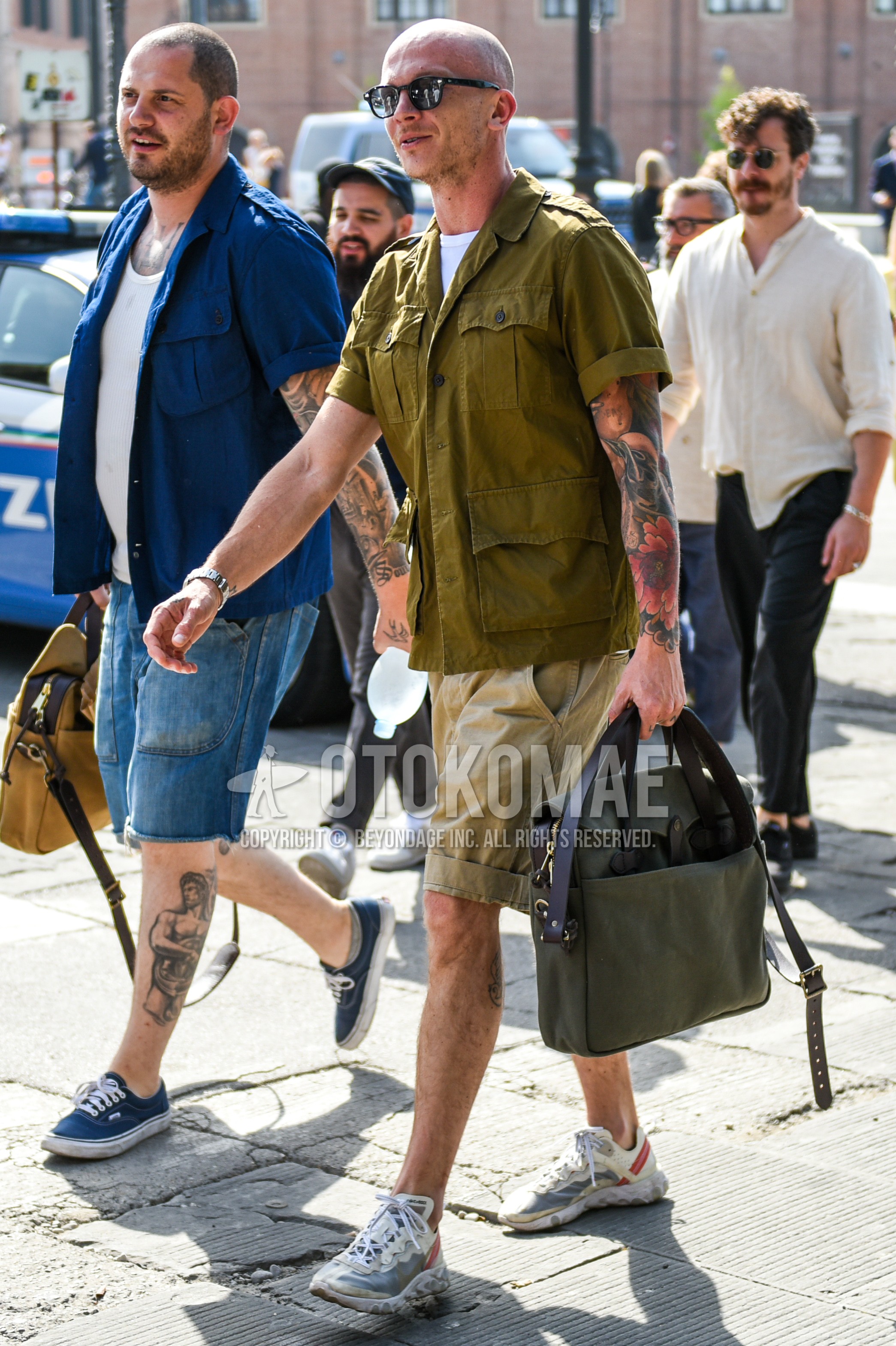 Men's summer outfit with black plain sunglasses, olive green plain M-65, white plain t-shirt, beige plain short pants, white low-cut sneakers, olive green plain shoulder bag.