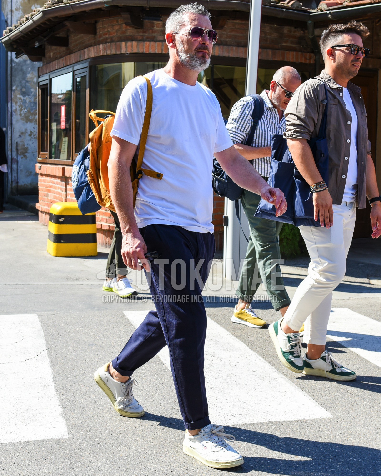 Men's spring summer outfit with silver plain sunglasses, white plain t-shirt, navy plain chinos, white low-cut sneakers, yellow plain backpack.