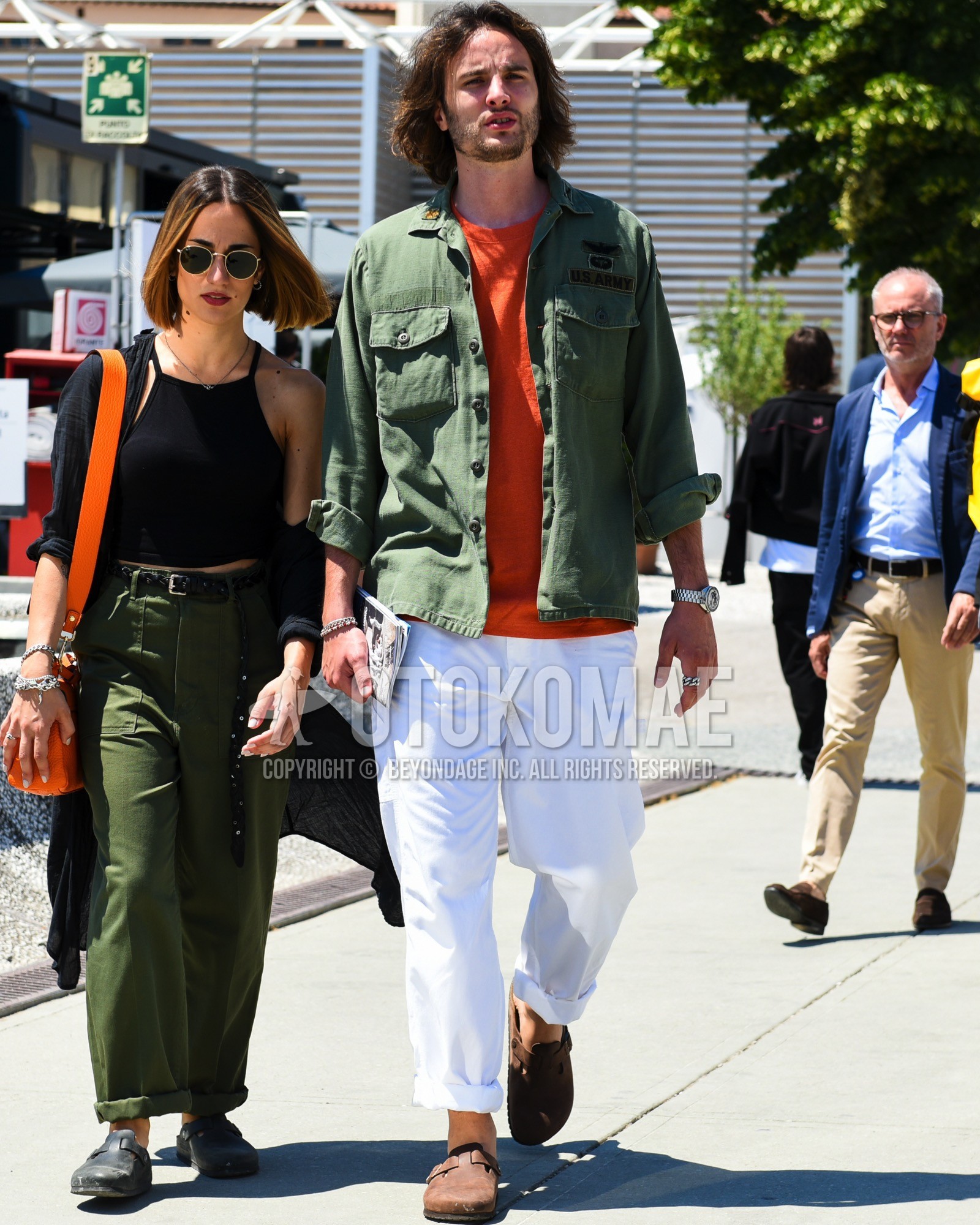 Men's spring summer outfit with olive green plain shirt jacket, olive green plain military jacket, orange plain t-shirt, white plain cotton pants, brown leather sandals.