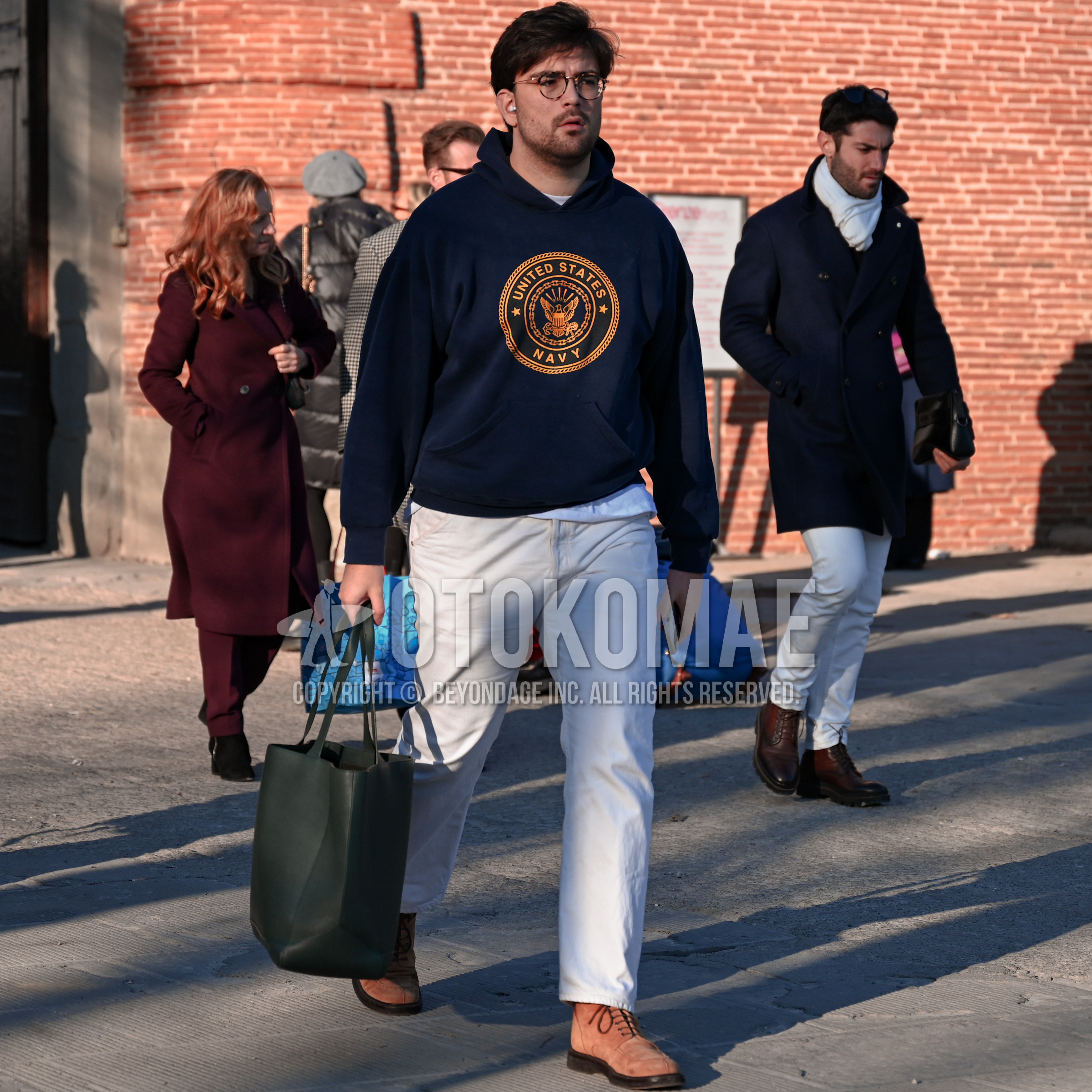 Men's autumn outfit with brown tortoiseshell glasses, navy deca logo hoodie, white plain long sleeve t-shirt, white plain cotton pants, beige suede shoes leather shoes, olive green plain tote bag.