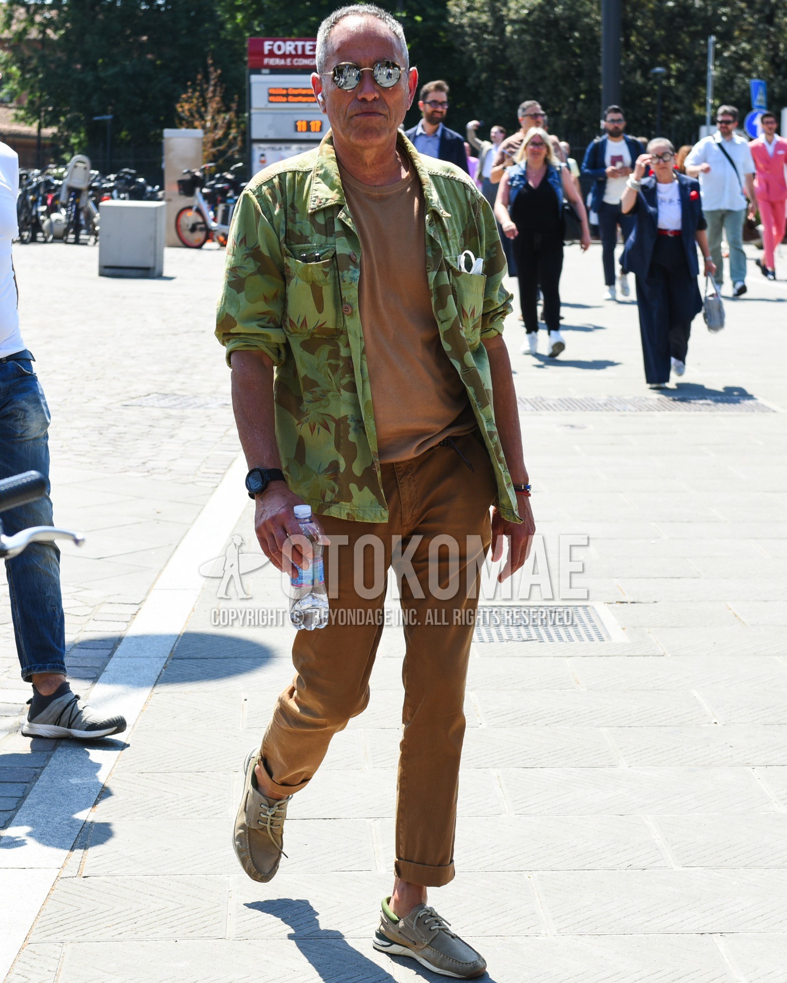 Men's spring summer outfit with brown tortoiseshell sunglasses, olive green camouflage shirt jacket, brown plain t-shirt, brown plain chinos, beige moccasins/deck shoes leather shoes.
