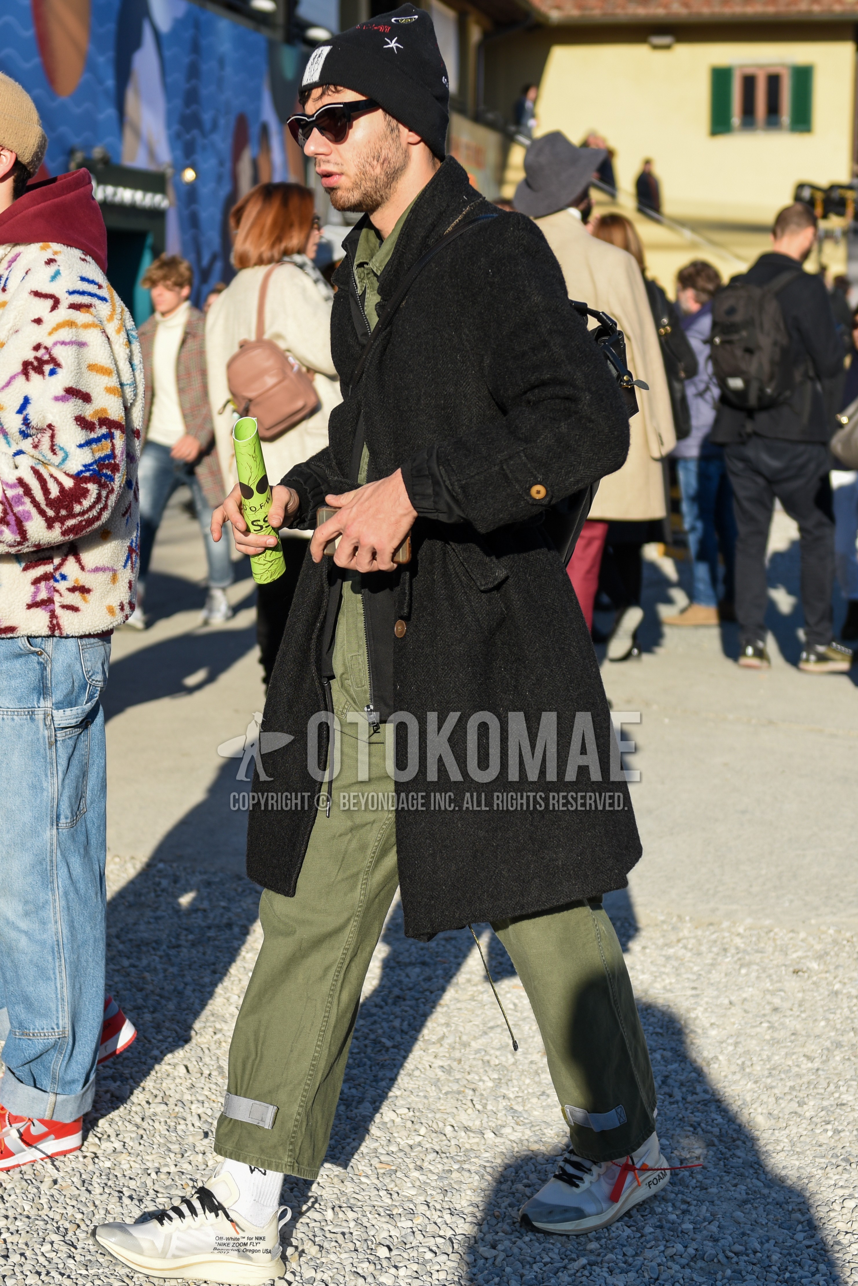 Men's autumn winter outfit with black plain knit cap, black plain sunglasses, dark gray herringbone stenkarrer coat, olive green plain jumpsuit, white plain socks, white low-cut sneakers, black plain backpack.