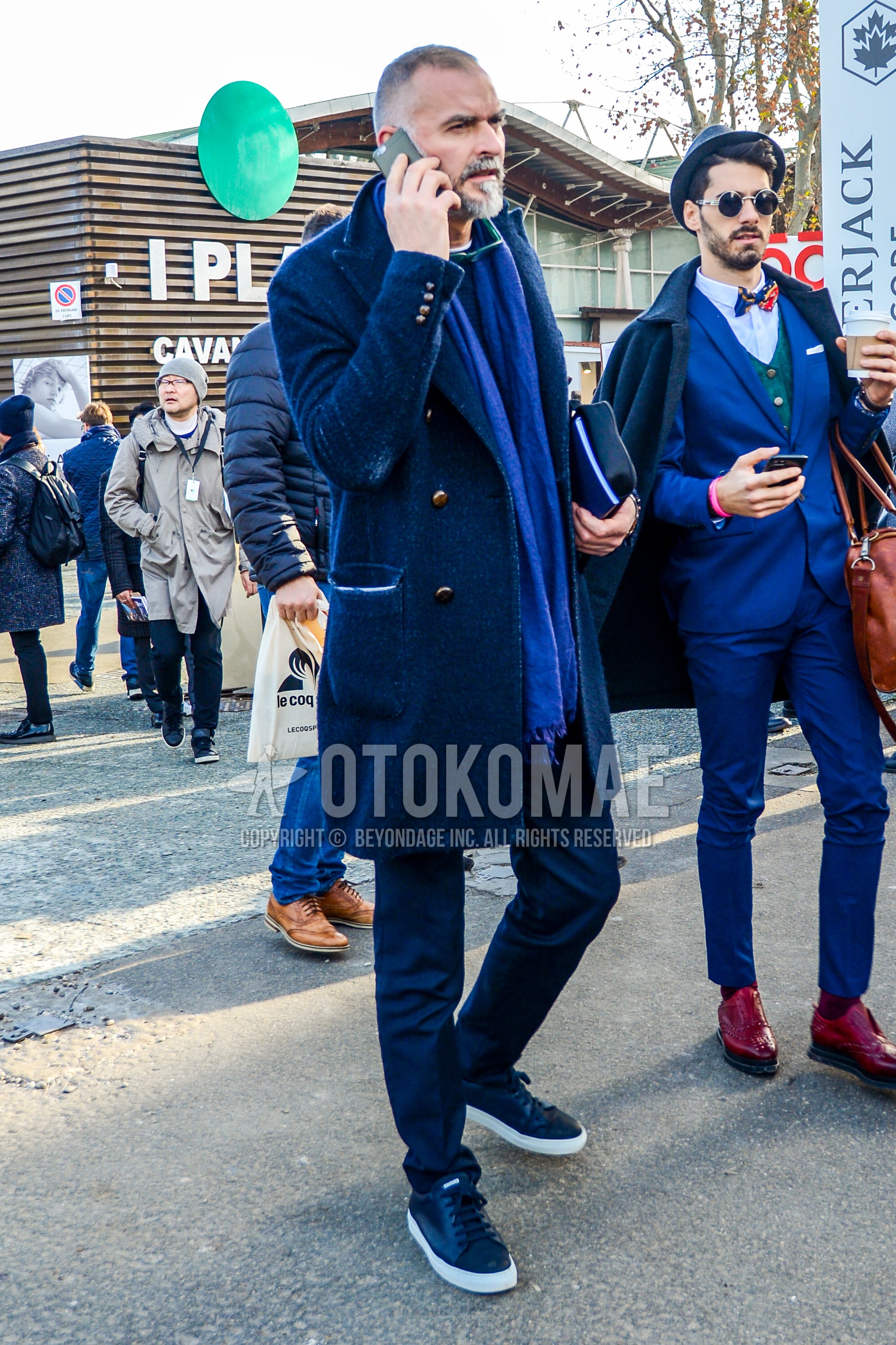 Men's autumn winter outfit with blue plain scarf, navy plain chester coat, gray plain slacks, black low-cut sneakers.