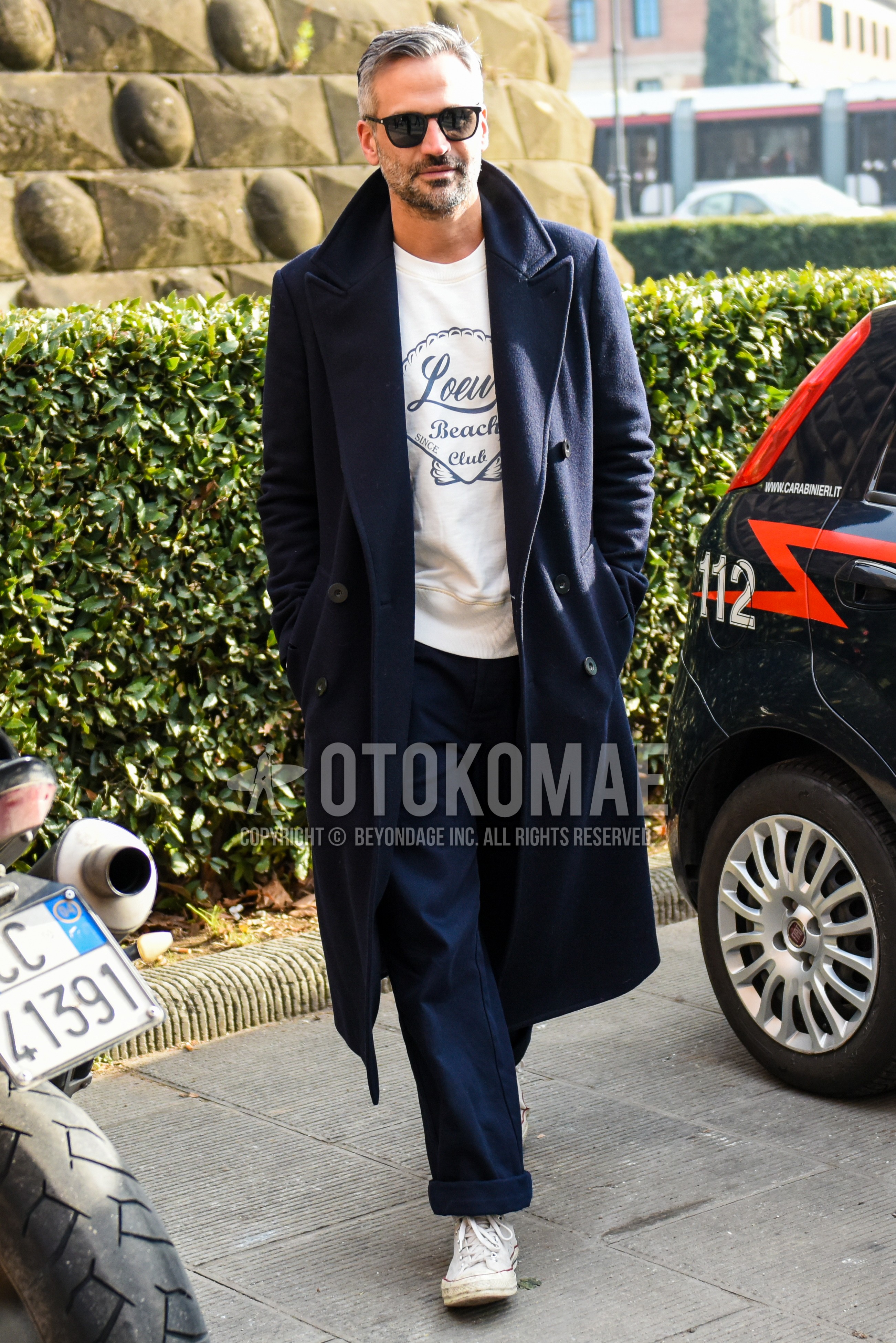 Men's autumn winter outfit with black plain sunglasses, navy plain chester coat, white navy graphic sweatshirt, navy plain chinos, white high-cut sneakers.