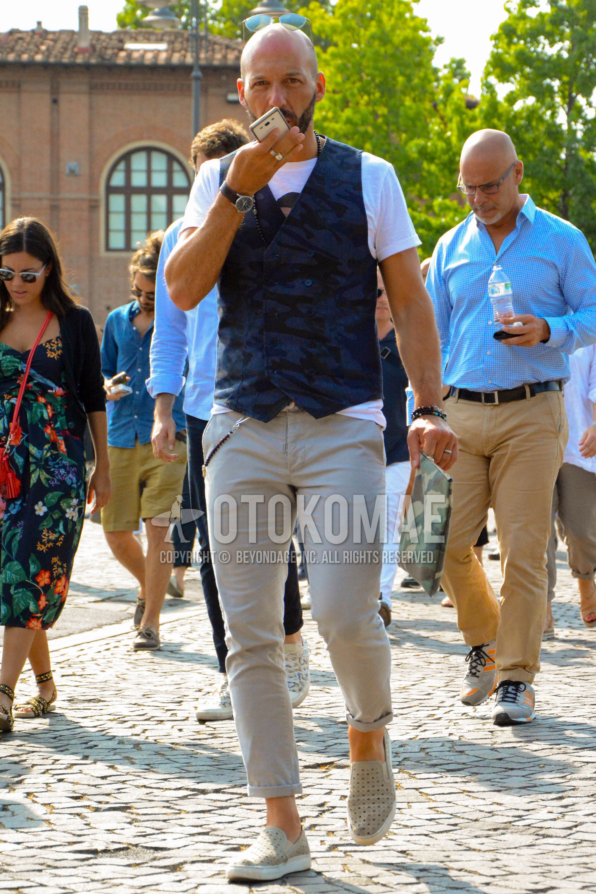 Men's spring summer outfit with white graphic t-shirt, blue camouflage gilet, beige plain chinos, beige slip-on sneakers.