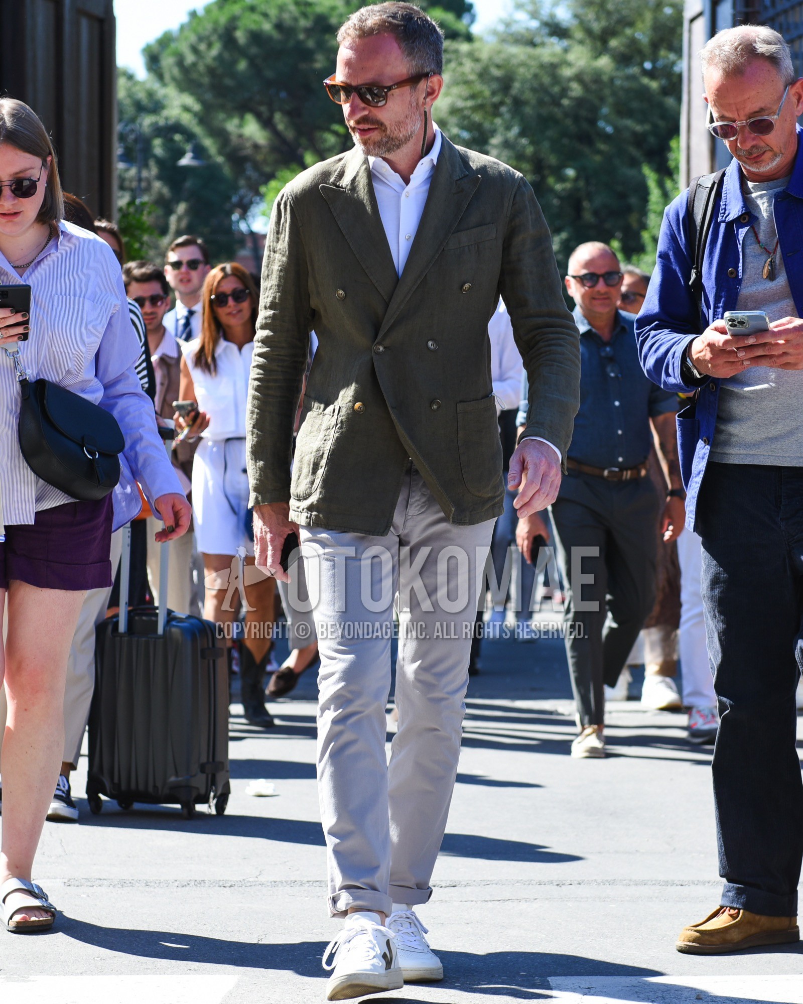 Men's spring summer outfit with brown plain sunglasses, olive green plain tailored jacket, white plain shirt, gray plain chinos, white low-cut sneakers.