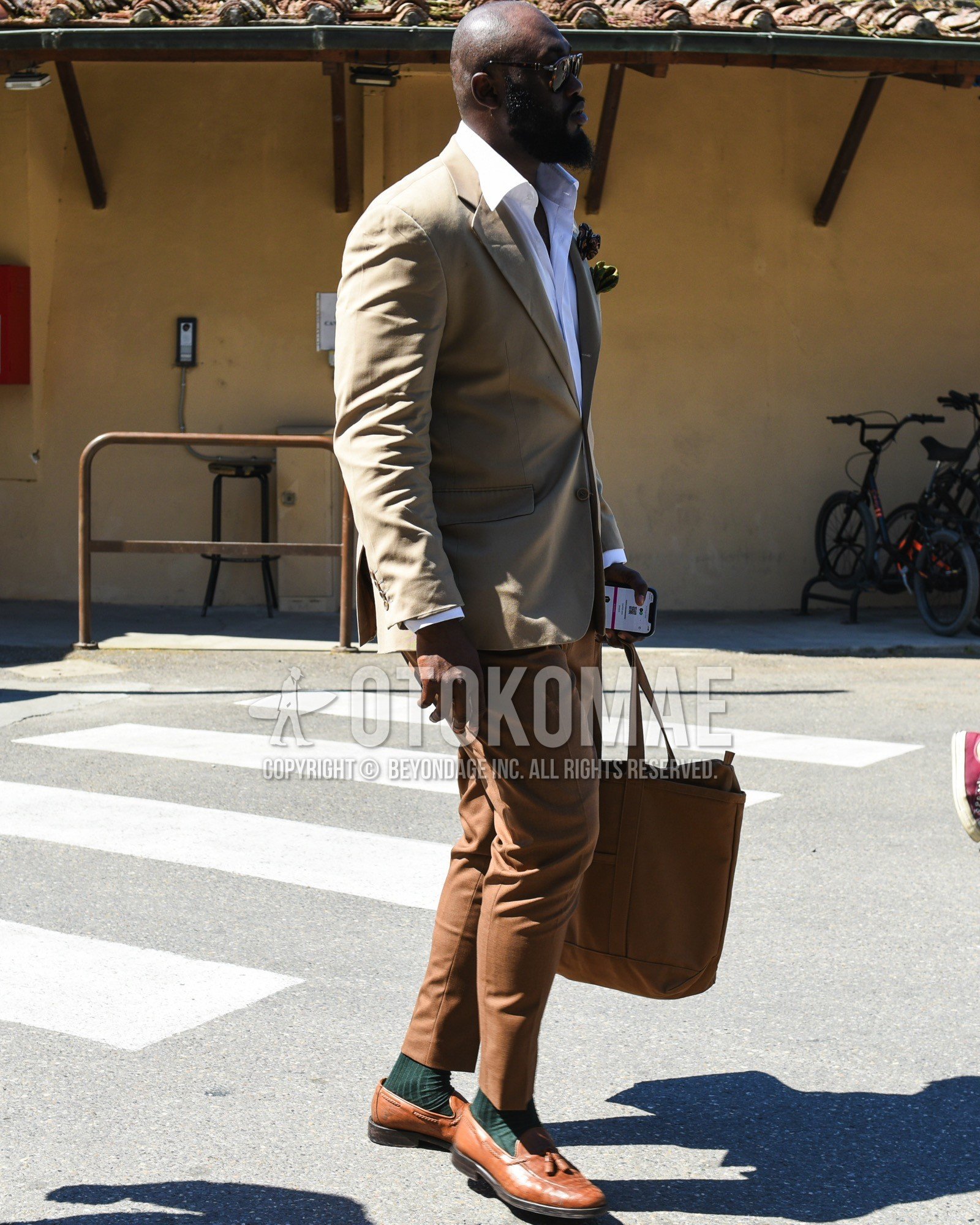 Men's spring summer outfit with brown tortoiseshell sunglasses, beige plain tailored jacket, white plain shirt, brown plain slacks, green plain socks, brown tassel loafers leather shoes, brown plain tote bag.