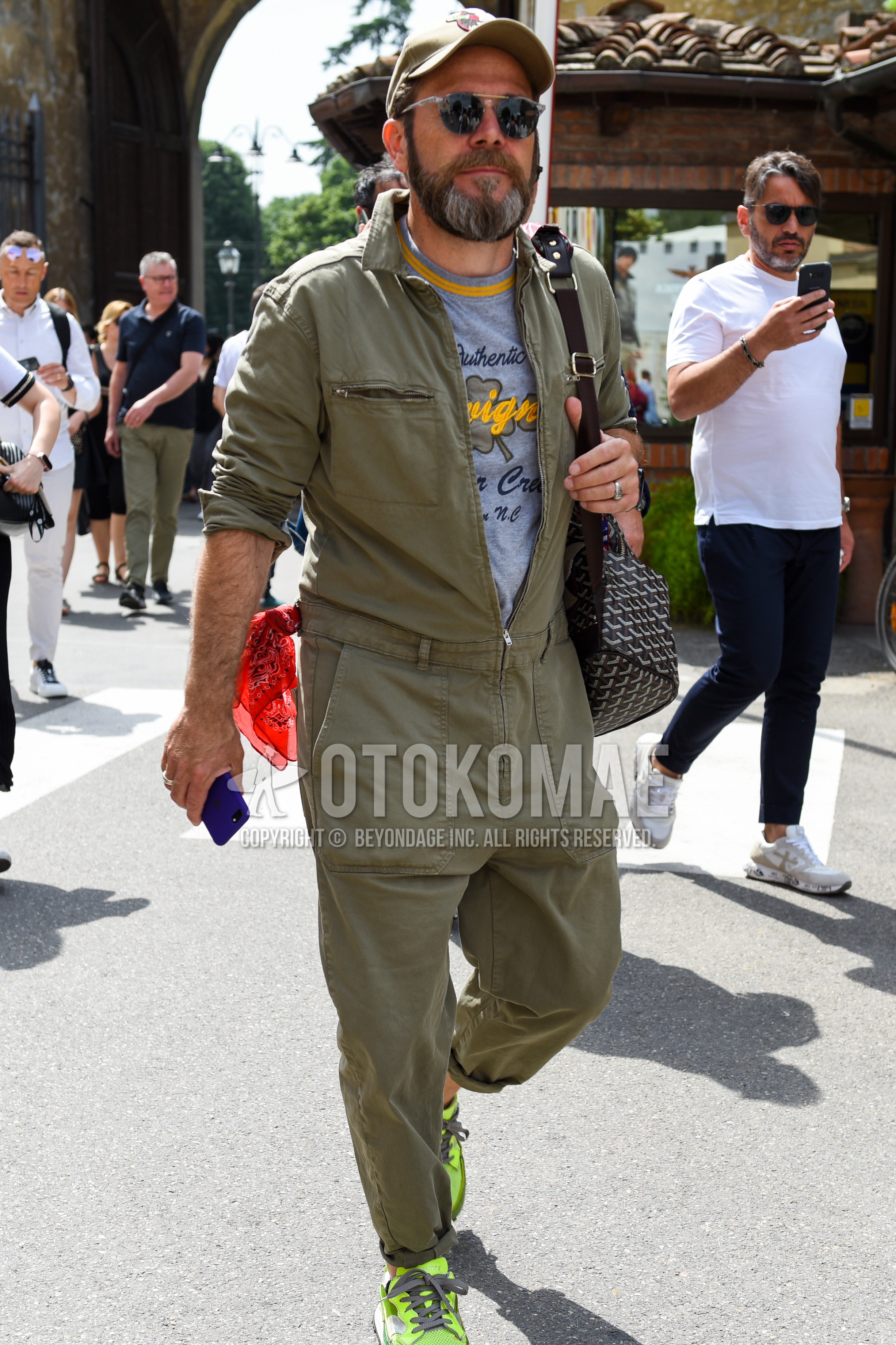 Men's spring autumn outfit with olive green one point baseball cap, gold black plain sunglasses, olive green plain jumpsuit, gray graphic t-shirt, yellow low-cut sneakers, brown gray plain shoulder bag.