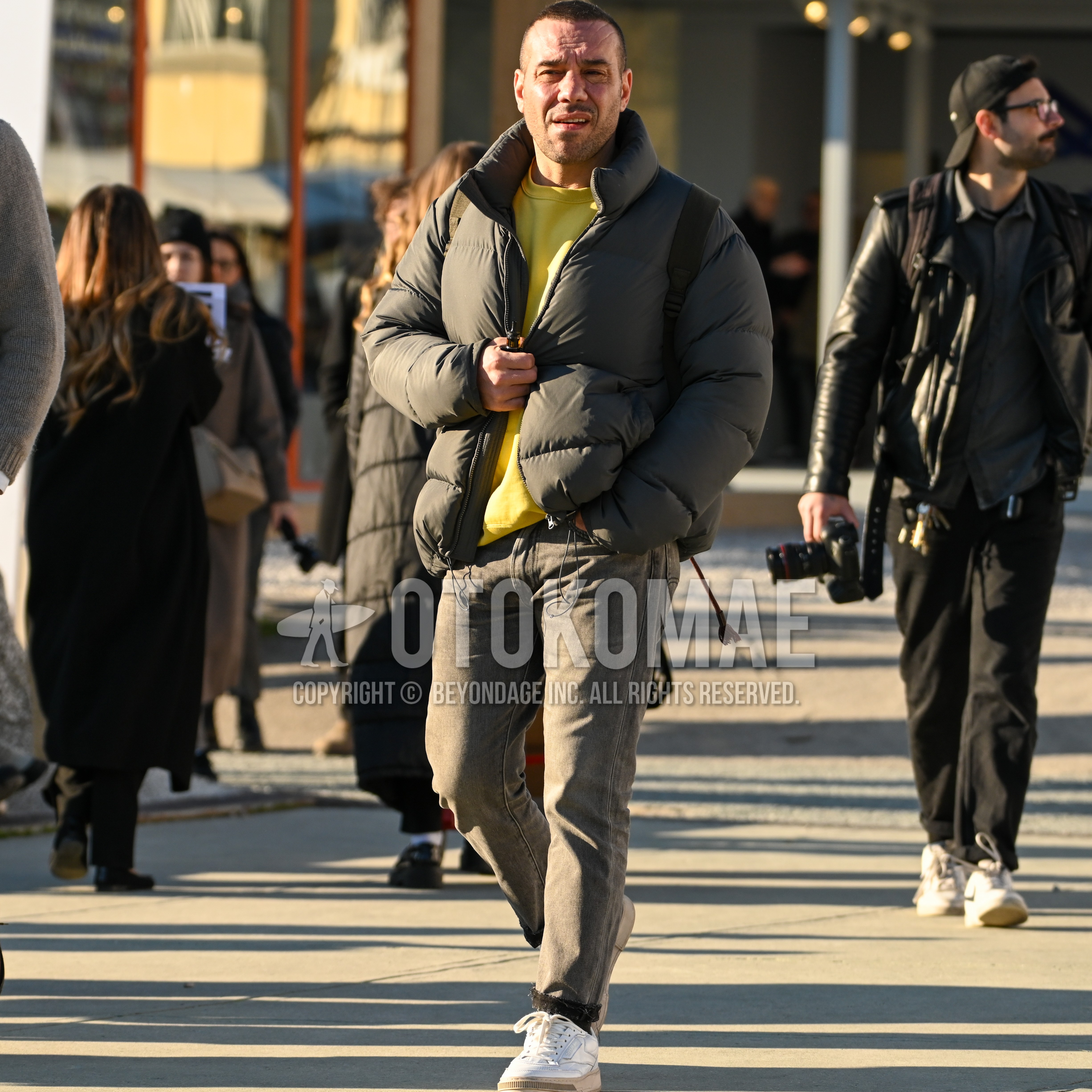 Men's autumn winter outfit with olive green plain down jacket, yellow plain sweatshirt, dark gray plain denim/jeans, white low-cut sneakers.