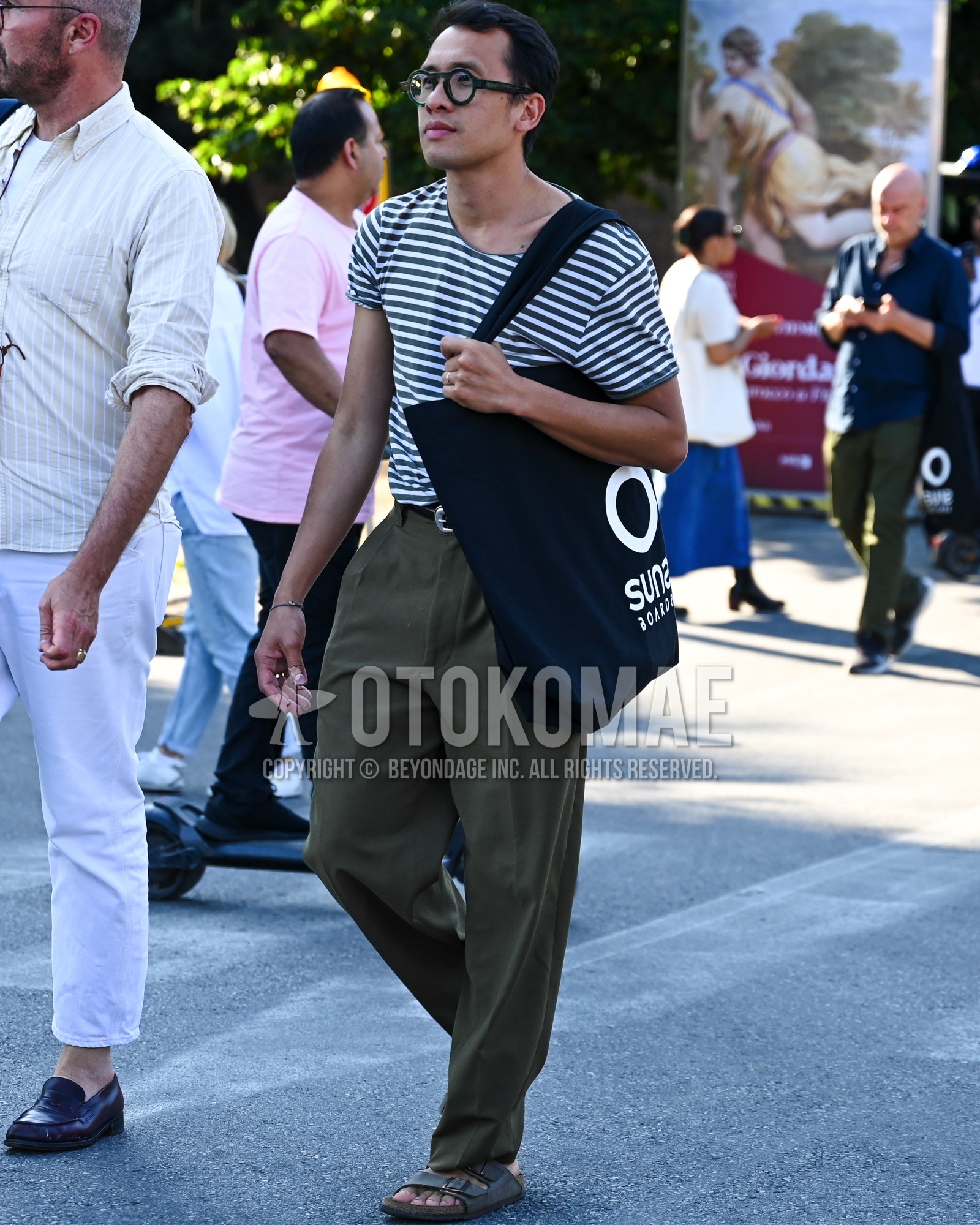 Men's spring summer outfit with clear plain sunglasses, white horizontal stripes t-shirt, olive green plain chinos, gray sport sandals, black deca logo tote bag.