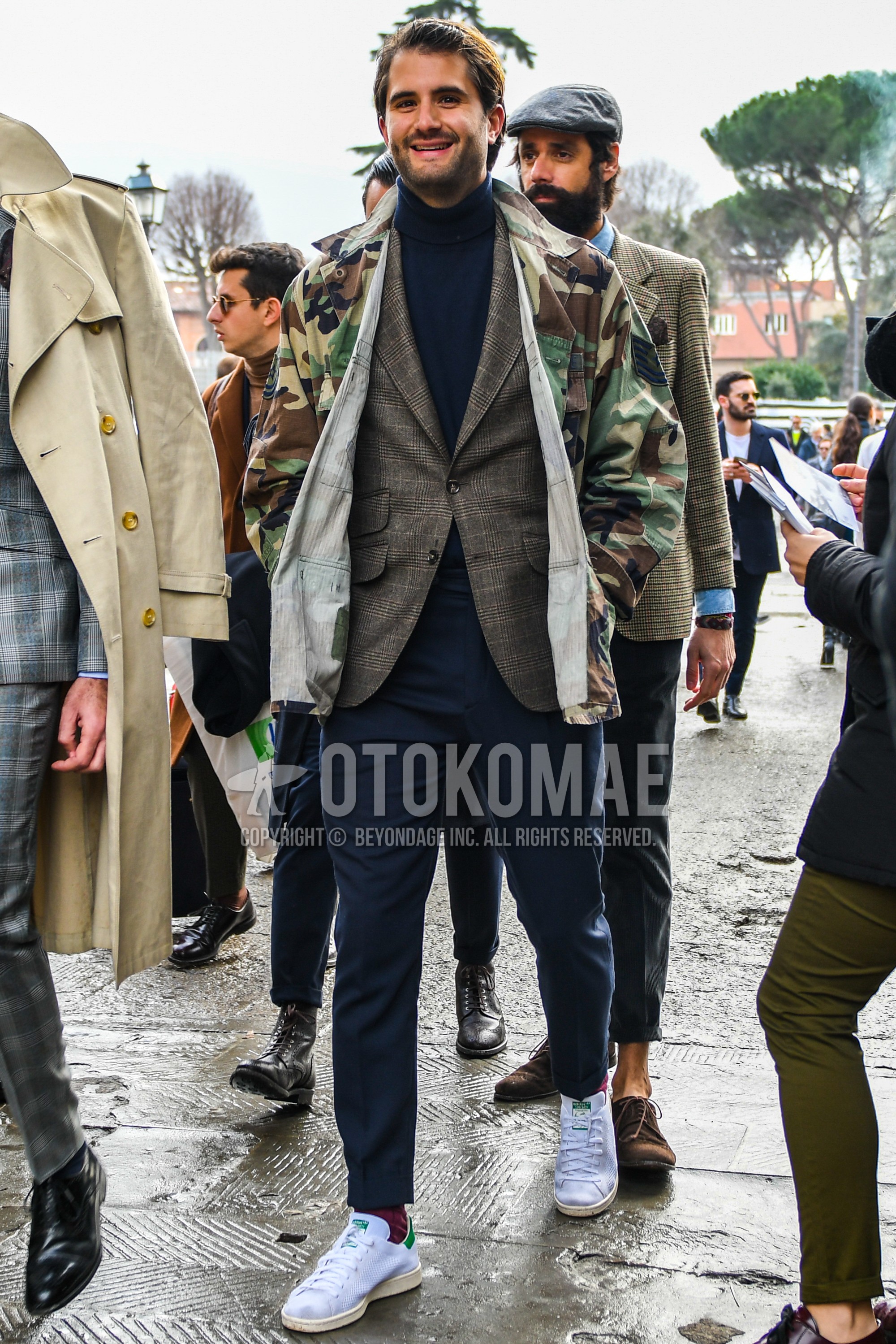 Men's winter outfit with multi-color camouflage safari jacket, brown check tailored jacket, navy plain turtleneck knit, navy plain slacks, red plain socks, white low-cut sneakers.