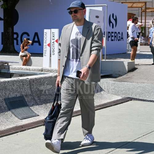 ブルーキャップのジェットキャップ、ブラウンべっ甲のサングラス、白グラフィックのTシャツ、白無地のソックス、白ローカットのスニーカー、ネイビー無地のトートバッグ、グレー無地のスーツを合わせた夏春のメンズコーデ・着こなし。