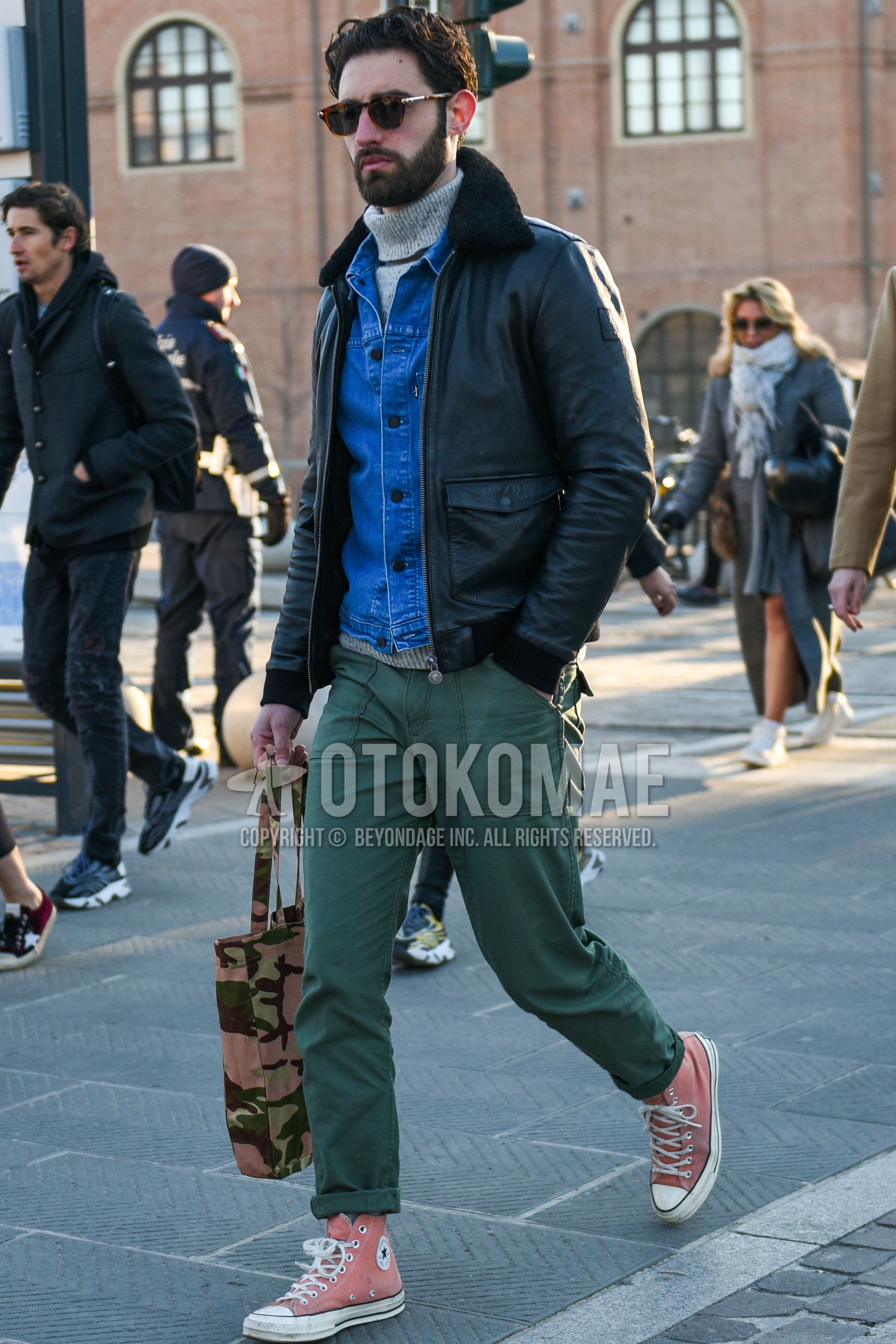 Men's autumn winter outfit with brown tortoiseshell sunglasses, black plain leather jacket, blue plain denim jacket, black plain military jacket, green plain turtleneck knit, olive green plain chinos, gray plain socks, pink low-cut sneakers, multi-color camouflage tote bag.