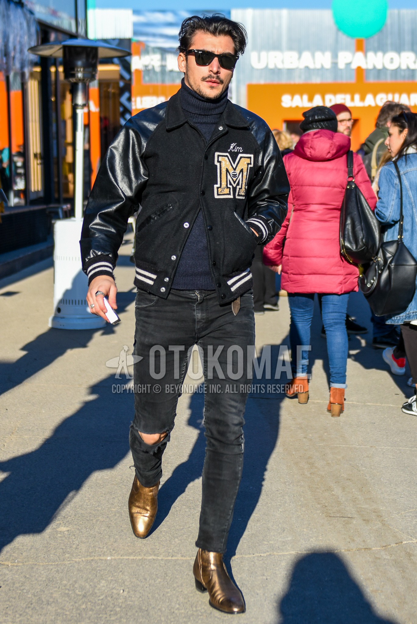 Men's autumn winter outfit with black plain sunglasses, black lettered stadium jacket, dark gray plain turtleneck knit, black plain damaged jeans, gold side-gore boots.
