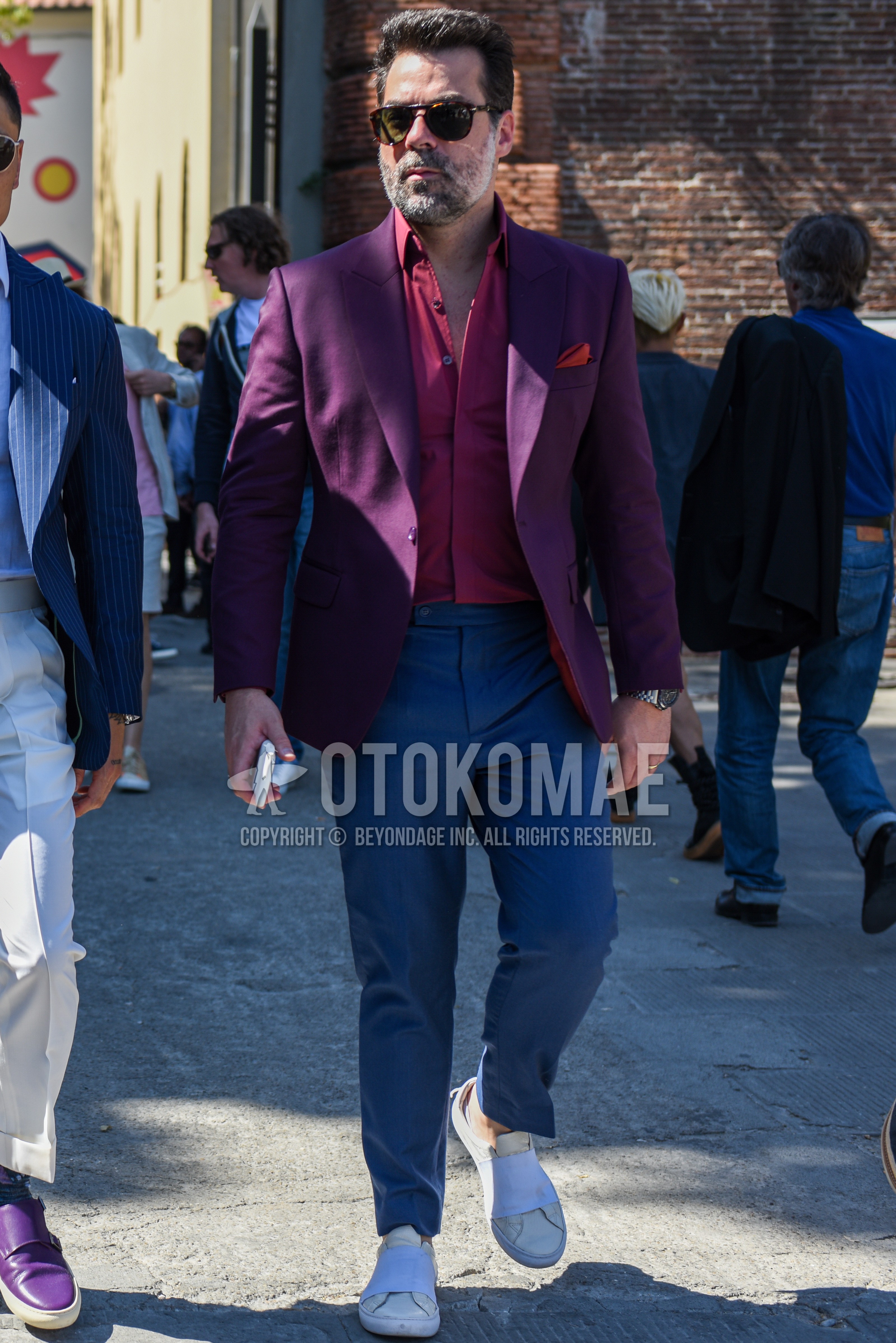 Men's spring summer autumn outfit with black plain sunglasses, purple plain tailored jacket, red plain shirt, blue plain beltless pants, white slip-on sneakers.