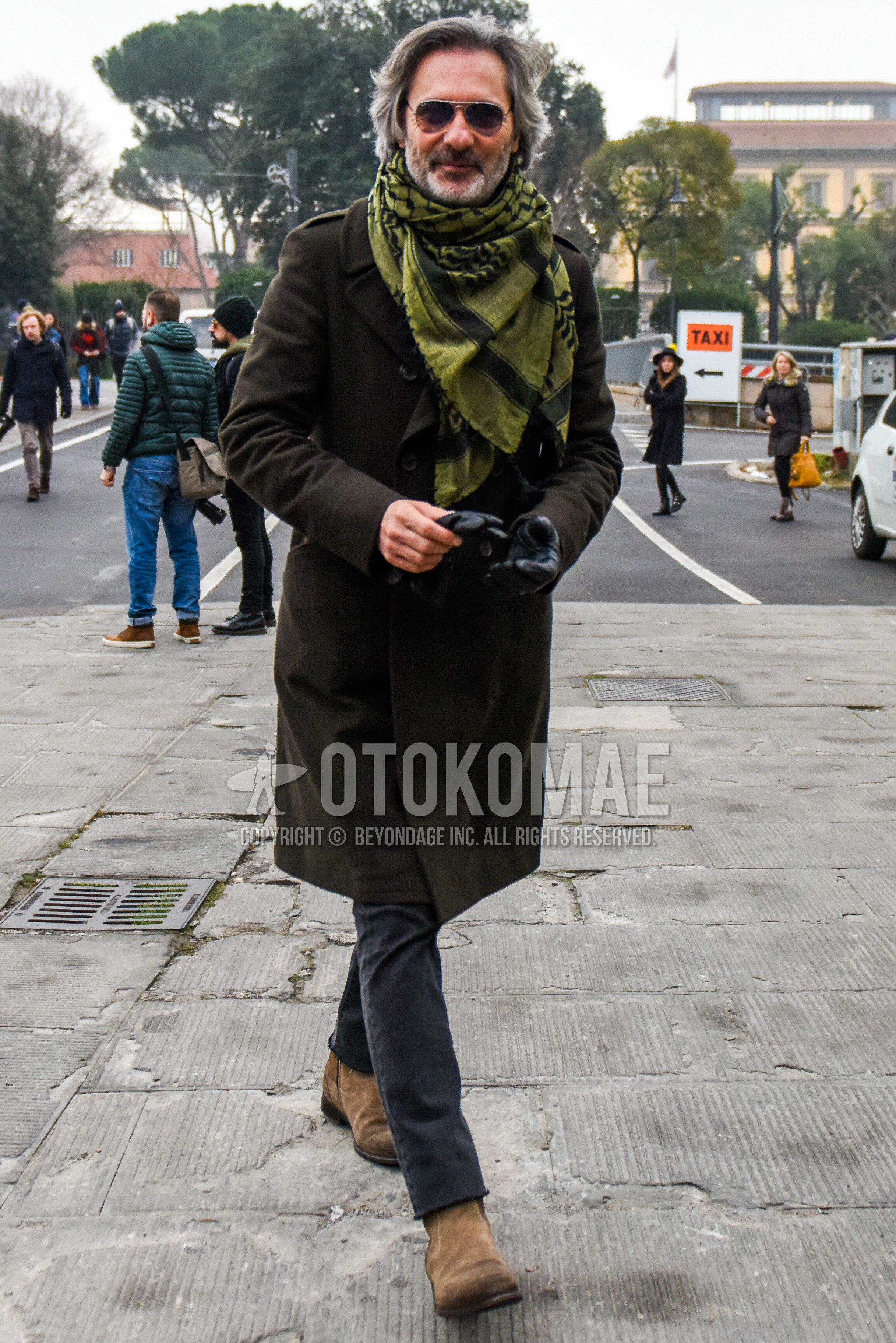 Men's winter outfit with silver plain sunglasses, olive green scarf scarf, olive green plain ulster coat, dark gray plain denim/jeans, beige side-gore boots.